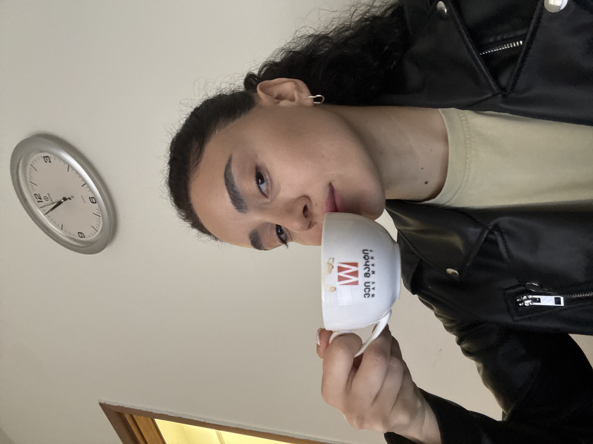 A person is holding a white coffee cup with Georgian text and logo of "Waymart" (likely a cafe chain) in a room with a clock on the wall. The clock reads approximately 1:33. The person is wearing a black leather jacket and a beige shirt. The location is likely in Tbilisi, Georgia, based on the Georgian text on the coffee cup.