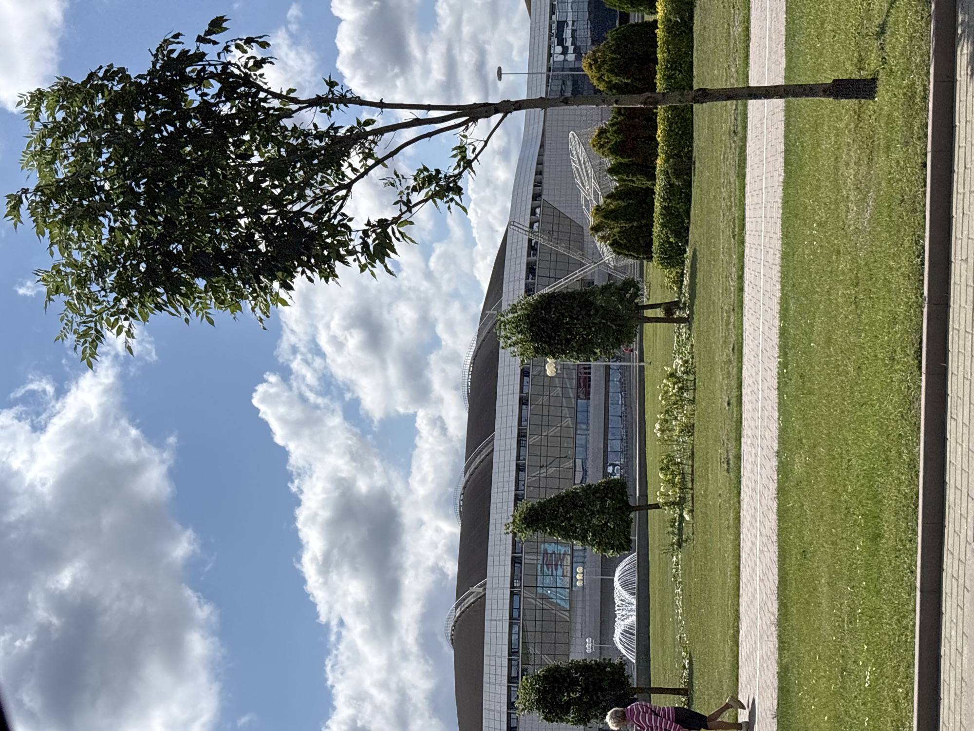 The image shows a woman walking on a paved path in front of a modern building with a curved roof. The building appears to be a stadium or arena, with large windows and a silver facade. In the foreground, there is a grassy lawn with several trees and a fountain. The sky is blue with scattered white clouds. The woman is wearing a pink and white striped shirt and dark shorts, and is walking from left to right. The environment is clean and well-maintained, suggesting a public space. The building's architecture is contemporary. This scene is located at the Minsk Arena, a multi-purpose sports and entertainment complex in Minsk, Belarus.