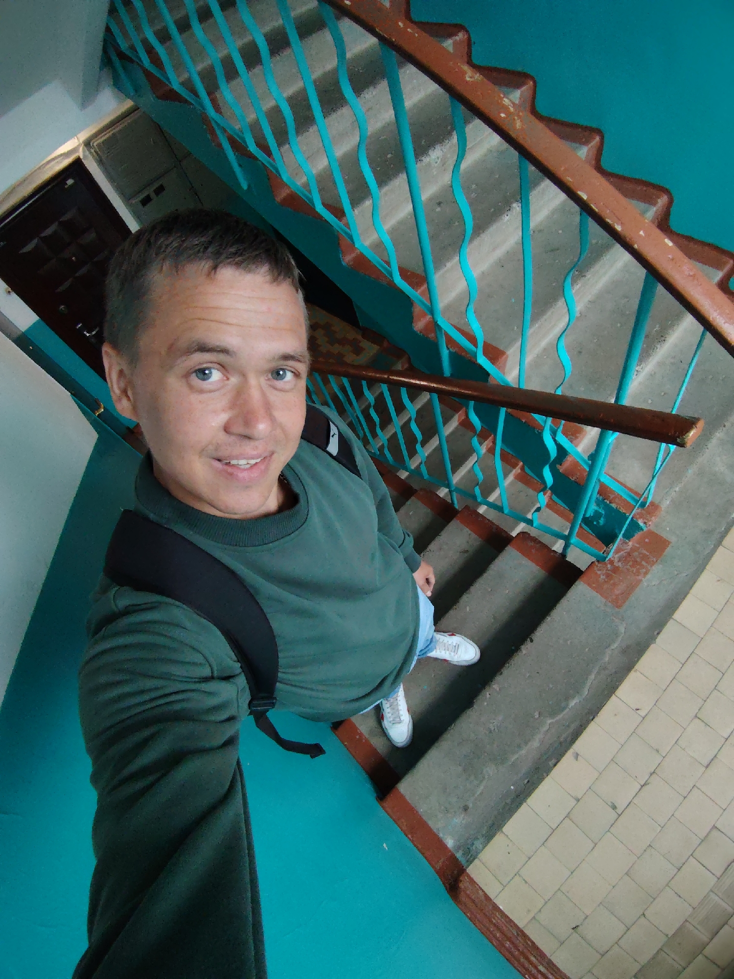 A male individual, appearing to be in his late 20s to early 40s with short light brown hair and blue eyes, is captured in a selfie. He is smiling slightly and looking directly at the camera. He wears a dark green long-sleeved sweatshirt, light blue jeans, and white sneakers with red accents. A black backpack is positioned on his back, with one strap visible over his left shoulder (viewer's right). The scene is an interior stairwell within a building in Chaikovsky, Russia. The man is positioned on a concrete step of the staircase. The surrounding walls are painted a vibrant turquoise blue, with a vertical white section visible to the left. The staircase features concrete steps, some exhibiting worn brown risers. A brown handrail, showing signs of wear, runs alongside the staircase, supported by decorative, wavy turquoise blue metal railings. A tiled floor composed of light beige square tiles is visible in the lower right corner. A dark brown door is partially visible in the upper left background.