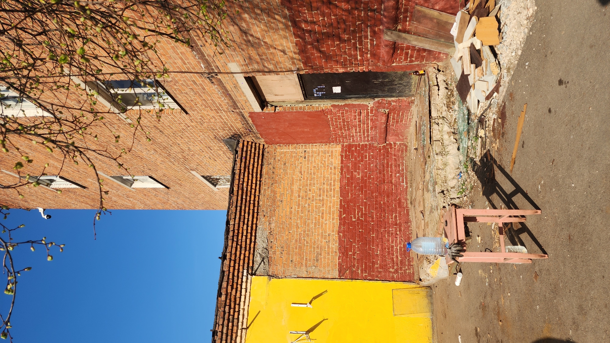 The image depicts an exterior shot of a building, potentially located in Zelenograd, Russia, given the query context. The building is constructed primarily of brick with sections painted in yellow, red, and orange. A black doorway is visible, partially covered with wood. Debris is scattered along the base of the building where it meets the paved ground. A small, weathered pink stool with a water bottle is situated on the pavement. The sky is a clear blue, and tree branches are visible on the upper left side of the frame.