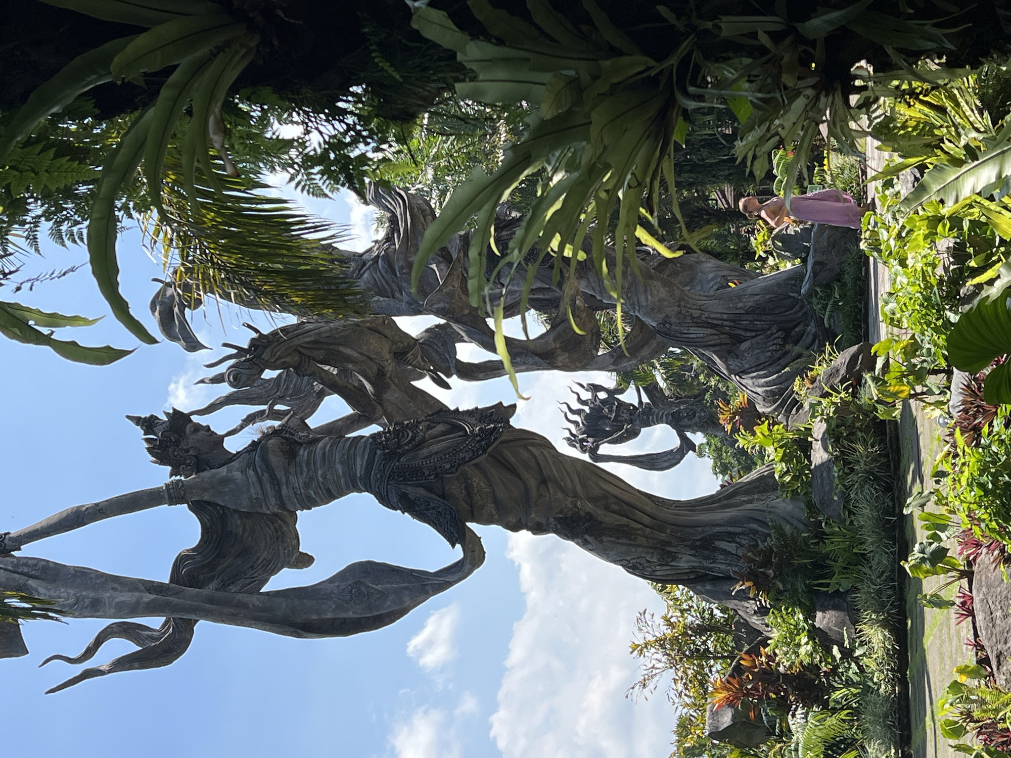 The image shows statues at the "Tirta Empul Temple" in Kedewatan, Ubud, Gianyar Regency, Bali, Indonesia. The gray, stone statues are adorned with traditional Balinese decorations and are surrounded by lush tropical vegetation. A woman in a pink dress stands near one of the statues. The sky is blue with scattered clouds.