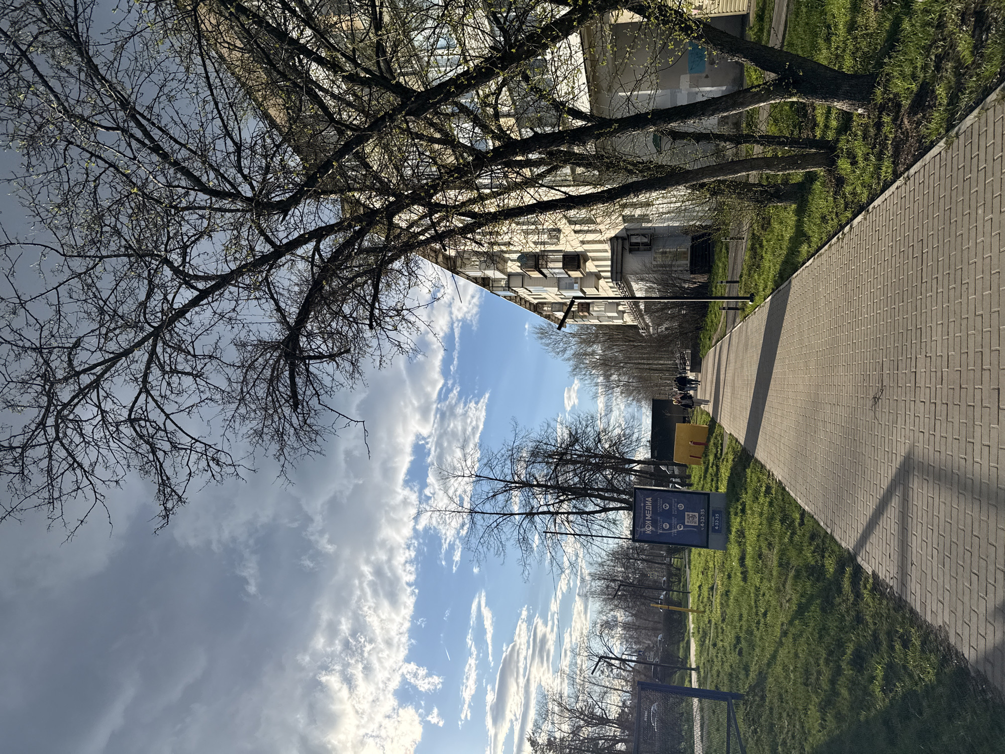 The image depicts an urban outdoor scene, likely a street or park path adjacent to residential buildings, captured in early spring. The photograph is oriented vertically, but for clarity, the description assumes a corrected horizontal view.
In the foreground, a paved pathway made of brick-like pavers extends from the bottom right towards the center and recedes into the distance. To the left of this path is a strip of vibrant green grass, which separates the path from a row of trees and buildings. The trees, characteristic of early spring, have mostly bare branches, though some show nascent green buds.
In the midground, beyond the first line of grass and trees on the left, stands a multi-story apartment building with numerous windows and balconies. On the right side of the pathway, there's another expanse of grass with more trees and a prominent blue signpost. Further down the path, small groups of people can be seen walking, suggesting it's a public area. A dark mesh fence or bin is visible in the lower right corner.
The sky occupies the upper portion of the frame, presenting a mix of blue patches and scattered white and grey clouds. The lighting suggests it is daytime, possibly late morning or afternoon, with visible shadows cast on the pathway and grass.
A notable detail is the blue signpost in the midground, which features Cyrillic text. The most prominent readable text is "КОИ МЕДИА" (KOI MEDIA), along with a QR code and what appears to be a phone number: "14-32-35". Several small icons are also visible on the sign, possibly indicating services or social media platforms. The overall scene conveys a quiet, everyday atmosphere in a residential or mixed-use urban environment.