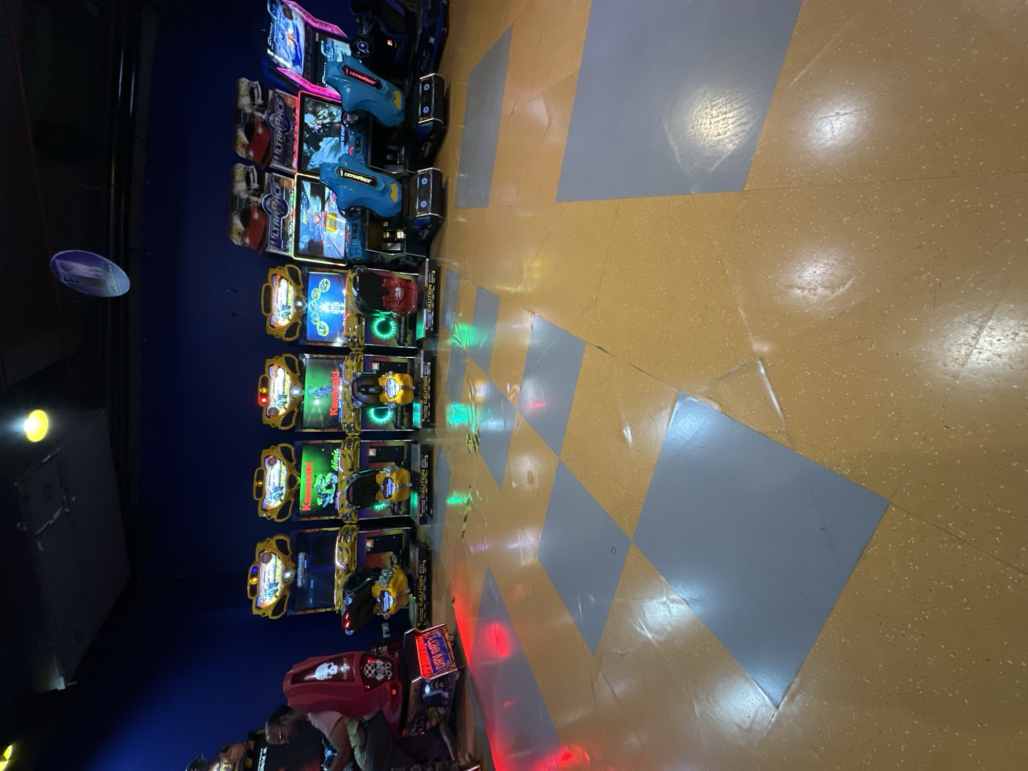 The image depicts a row of arcade games in an indoor setting, likely an entertainment center in Kalyan-Dombivli, India. The arcade games are arranged along a wall with blue paint. There are several games that appear to be motorcycle racing games, followed by a pair of blue gun-themed racing games near the end of the row. The floor has a diamond pattern with yellow and grey tiles. People are visible near the bottom of the frame.