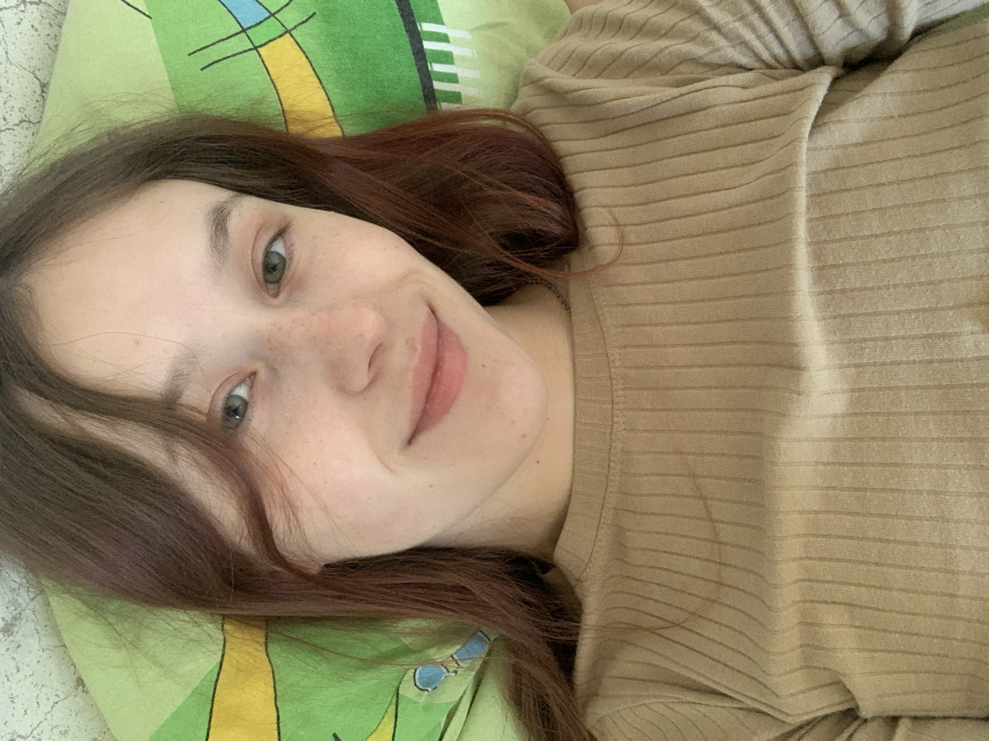 A young woman with reddish-brown hair lies on a green and white patterned pillow. She is wearing a tan, horizontally ribbed shirt. Her face is visible and she is smiling slightly. The location is unknown.