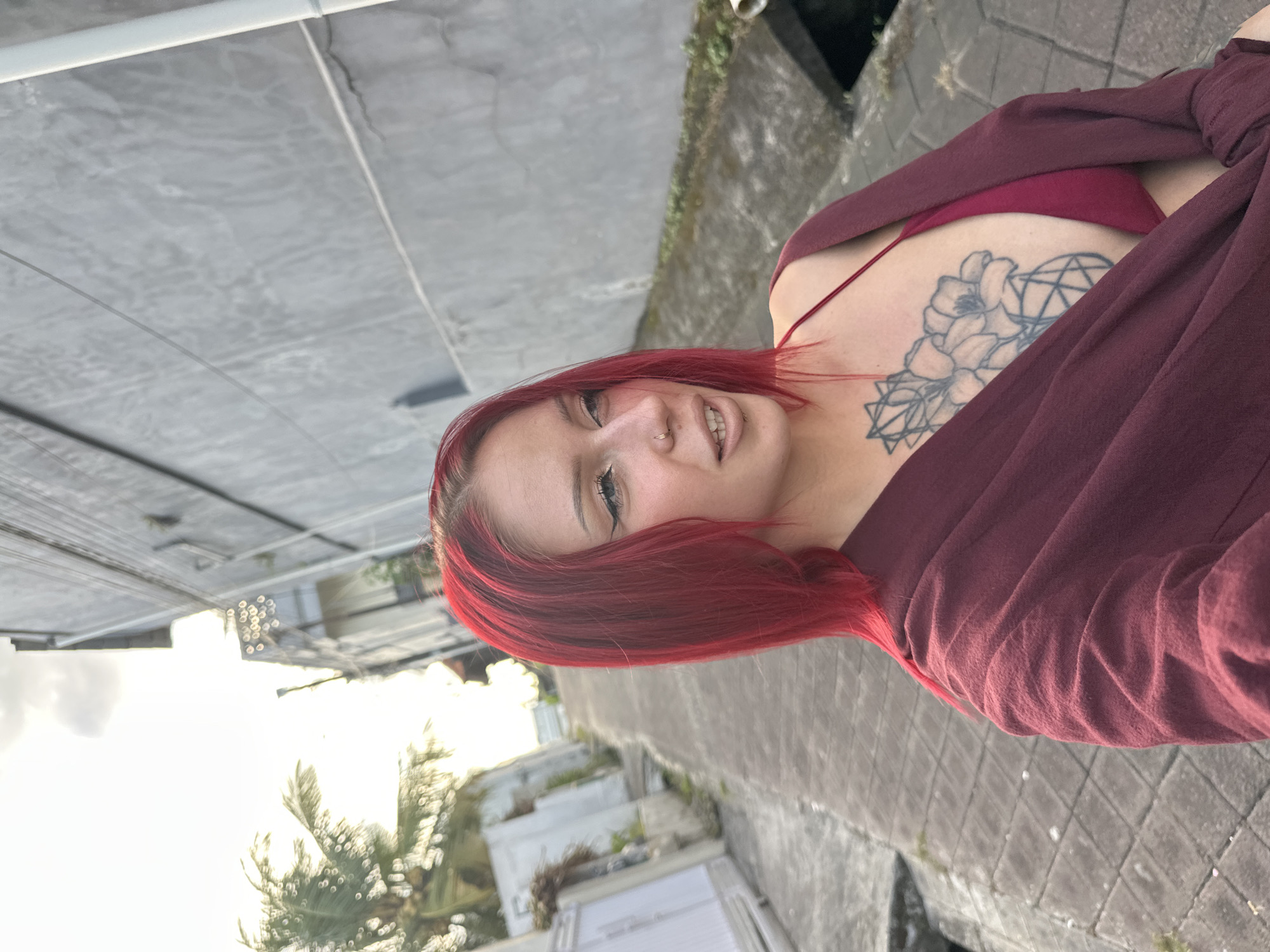 The image depicts a woman with red hair and a nose ring standing outside. She is wearing a maroon top and has a tattoo on her left shoulder. The background suggests an urban environment, with buildings, foliage, and paved areas visible. Given the context, it appears to be located in North Kuta, Indonesia.