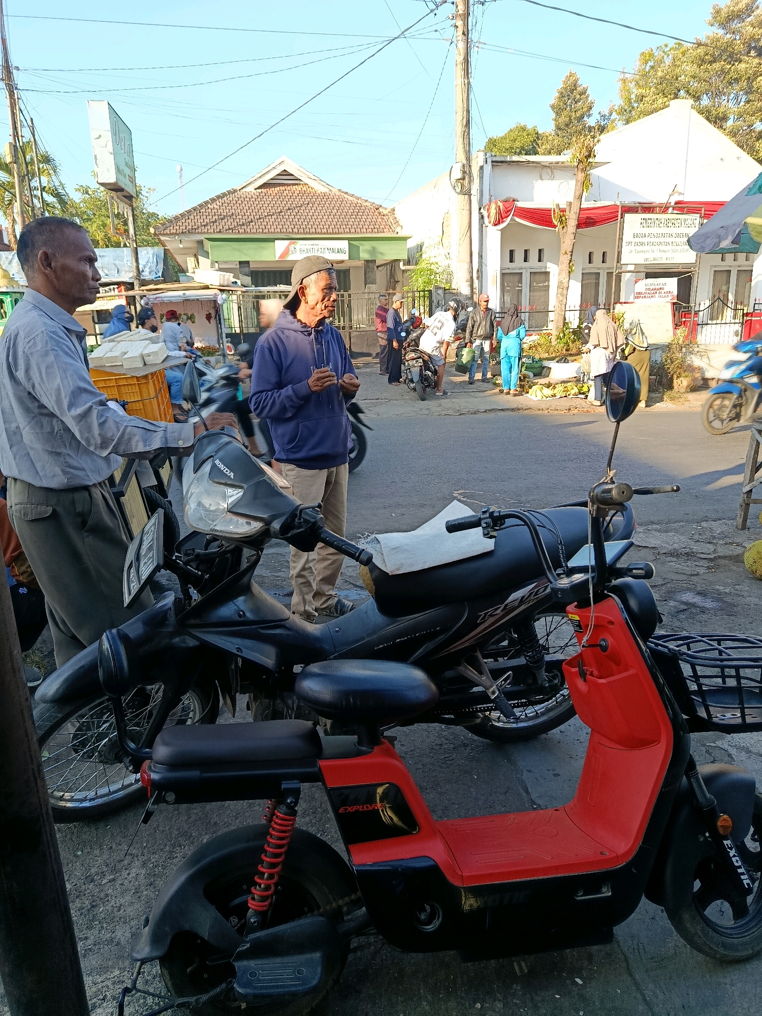 The image shows an outdoor scene in Bululawang, Indonesia, likely a street market or a government office entrance. In the foreground, two men are standing next to parked motorcycles. The man on the left, wearing a light blue collared shirt and dark pants, is facing to his right, looking towards a yellow crate filled with white rectangular objects, possibly tofu. The man on the right, wearing a blue hooded sweatshirt and tan pants, with a black baseball cap, is facing to his left, holding something in his hands.
Behind them, several other people are visible, some gathered around stalls selling produce, primarily bananas. A white building with red and white banners is in the background, with signage indicating it is "UPT BADAN PENDAPATAN DAERAH" (Regional Revenue Agency) and "BULULAWANG." Another building to the left has a similar architectural style and signage. The street is paved, and utility poles with numerous wires are present. The lighting suggests it is daytime, possibly morning or late afternoon, given the long shadows. The environment appears to be a mix of commercial and administrative activity.