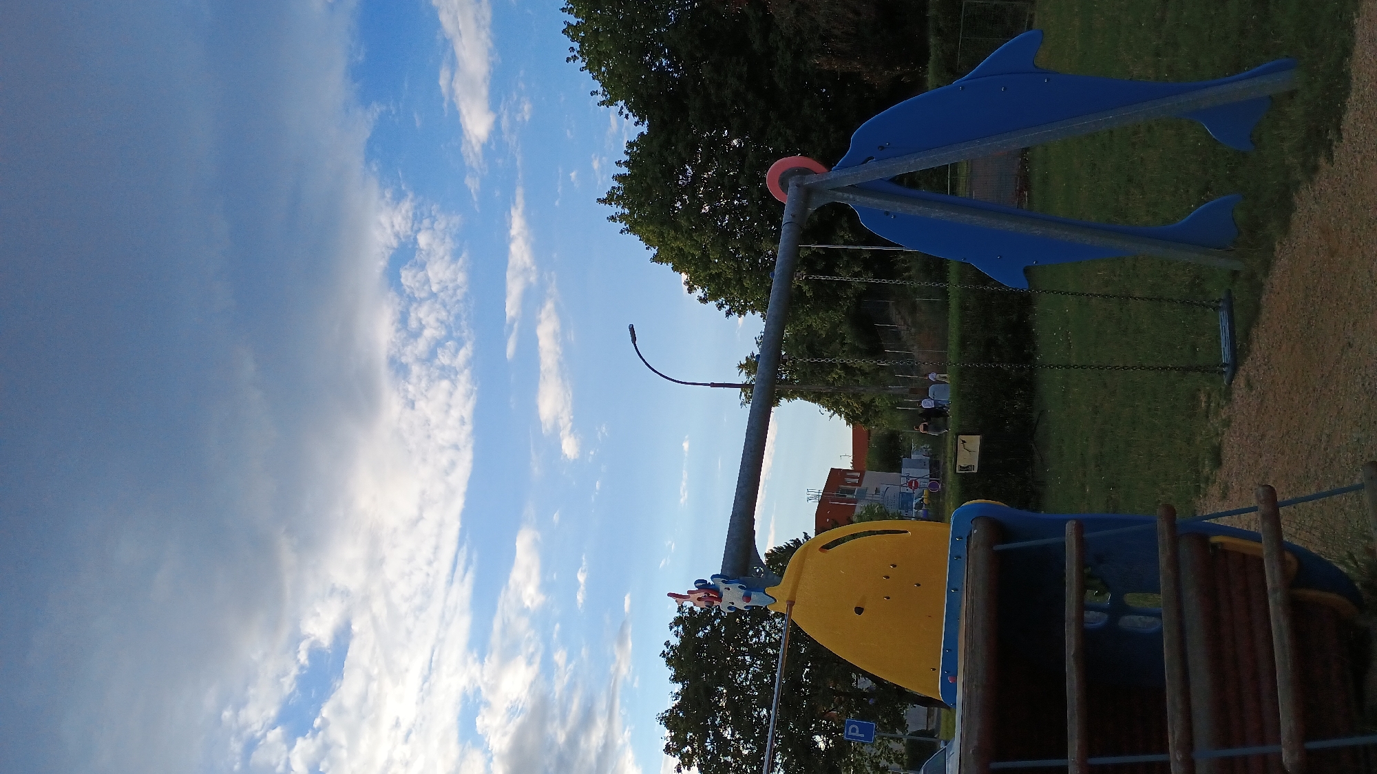 This image captures an outdoor scene at a playground in Sokolov, Czechia, likely during the late afternoon or early evening. The sky is a prominent feature, displaying a mix of blue sky and large, diffused clouds, with some areas appearing darker and more overcast.
In the foreground and midground, the playground equipment is visible. To the left, a portion of a blue and yellow play structure, possibly a boat or a slide, is partially cropped. It features a curved yellow section and horizontal wooden bars on its side. To the right, a swing set with two blue dolphin-shaped supports is prominent. A single swing, consisting of a dark blue seat suspended by chains, is hanging from the horizontal bar of the swing set.
The ground appears to be a mix of gravel and grass. In the background, trees with lush green foliage line the perimeter of the playground, some forming a dense barrier. Behind the trees, buildings with red roofs are visible, suggesting a residential or commercial area adjacent to the park. A street lamp with a curved pole stands near the center of the image, and a few people, appearing as small figures, are walking in the distance. A blue parking sign is also visible on the left side of the frame. The overall impression is a public recreational space under a dynamic sky.