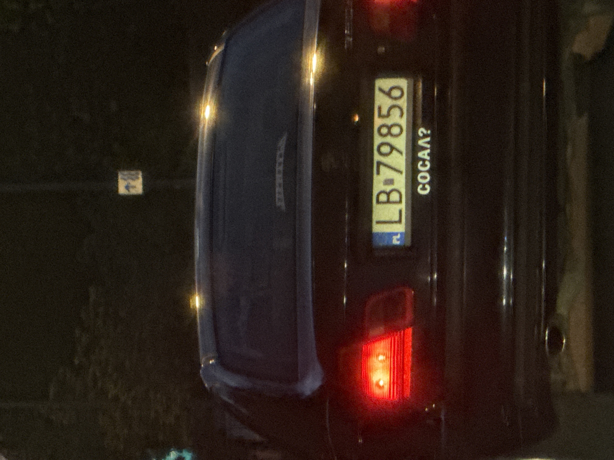 The image shows the rear of a black car parked on a street at night. The car has a license plate with the number "LB 79856" and the Polish (PL) identifier. Under the license plate, the word "COCAA?" is written. The car's taillight is illuminated. A sign is partially visible in the background on the left. The location is Warsaw, Poland, based on the PL license plate indicator.