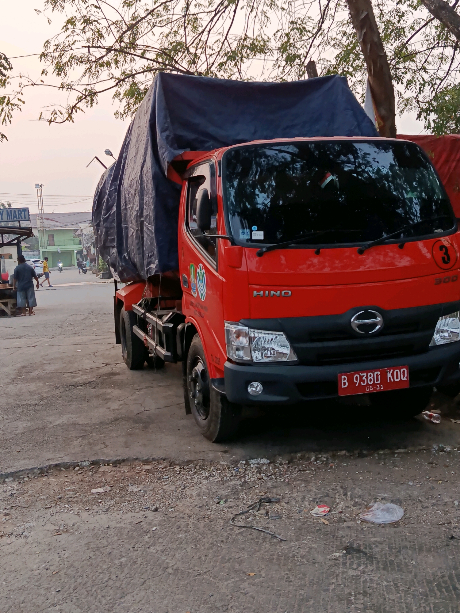 The image depicts a red Hino truck parked on a rough, asphalt surface with visible cracks and debris. The truck's cargo is covered by a large, dark blue tarp. The truck's front features a red license plate with white lettering that reads "B 9380 KOQ" and smaller text "05-31". A logo with three concentric circles and a stylized "3" is visible on the truck's hood. In the background, a sign that reads "Y MART" is partially visible, suggesting a commercial area. Several people are visible in the distance, including a person walking away from the viewer and two individuals near a building. Trees with sparse foliage are present, casting shadows. The sky appears overcast. The location is identified as a street in Bekasi, Indonesia.