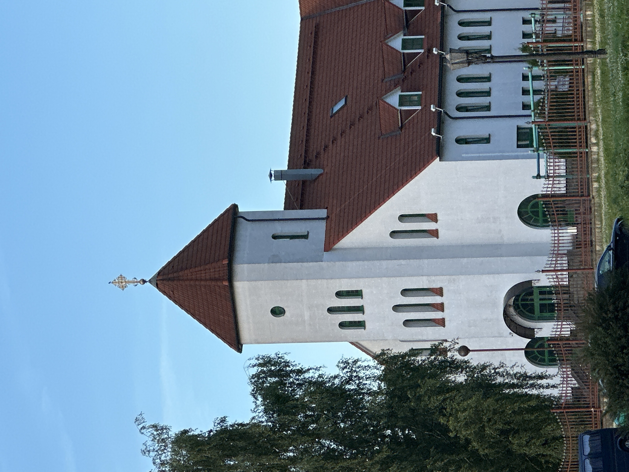 A white church building with a red tile roof stands under a clear blue sky. The building has a tall tower with a pointed roof, topped with a gold cross. The tower has several arched windows with green trim. The main section of the building also features arched windows with green trim and dormer windows on the roof. A large green tree with a drooping canopy is on the left side of the image, partially obscuring the church. A metal fence with a red railing runs along the front of the property, and a street lamp stands in front of the building. The ground in front of the fence is covered with green grass and some white flowers.