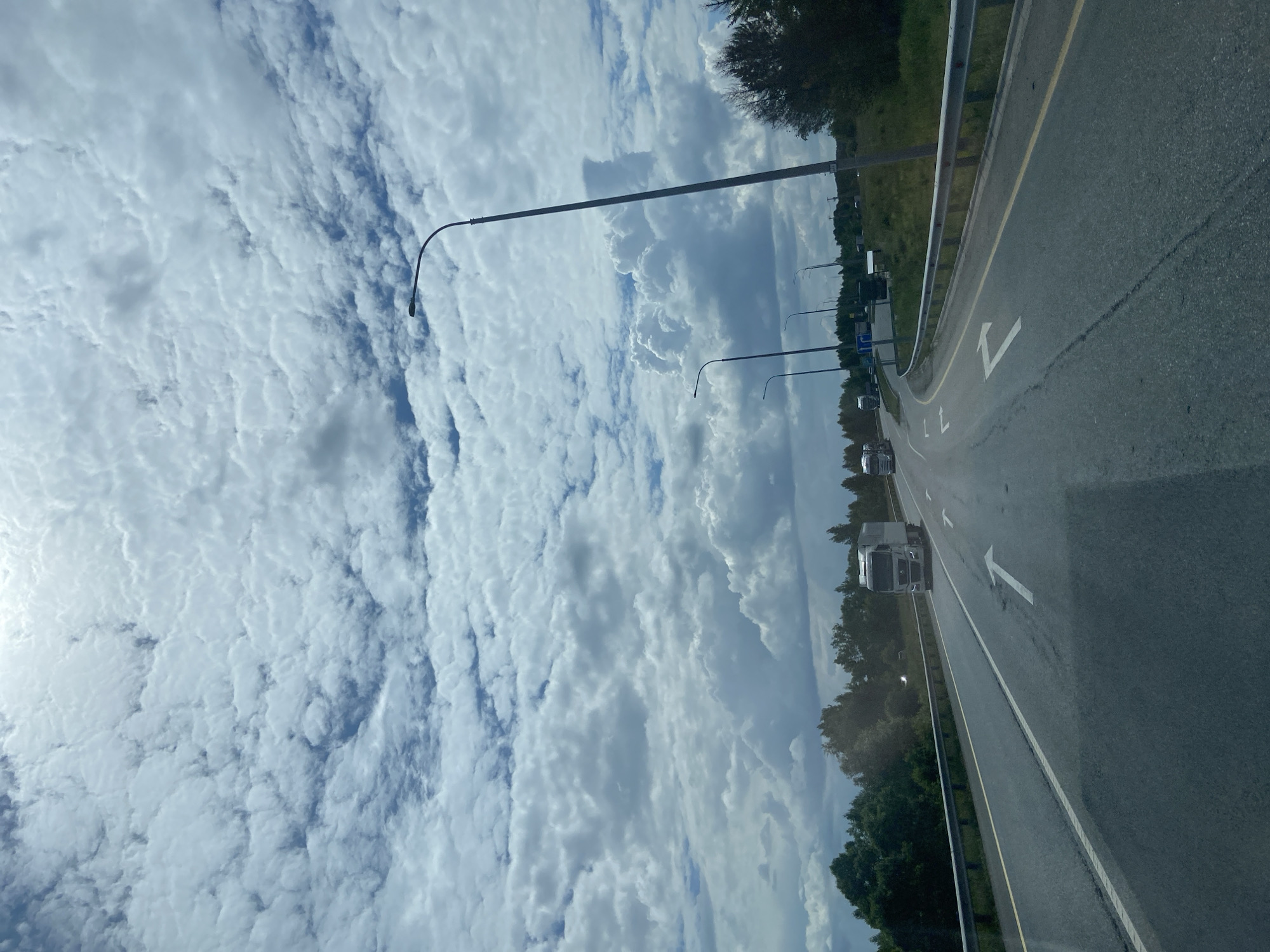 A view from a vehicle traveling on a highway with multiple lanes. The sky is filled with dramatic, white, puffy clouds against a light blue background. Streetlights with curved poles line the side of the road. On the right side of the road, there is a metal guardrail and grassy embankment with trees and foliage. In the distance, several trucks are visible on the highway. The road has white lane markings and directional arrows painted on its surface. The scene suggests a journey on a cloudy day in a semi-rural or suburban area. The location is identified as the city of Pervomaiskoye, Russia, implying it's a road within or near this city.