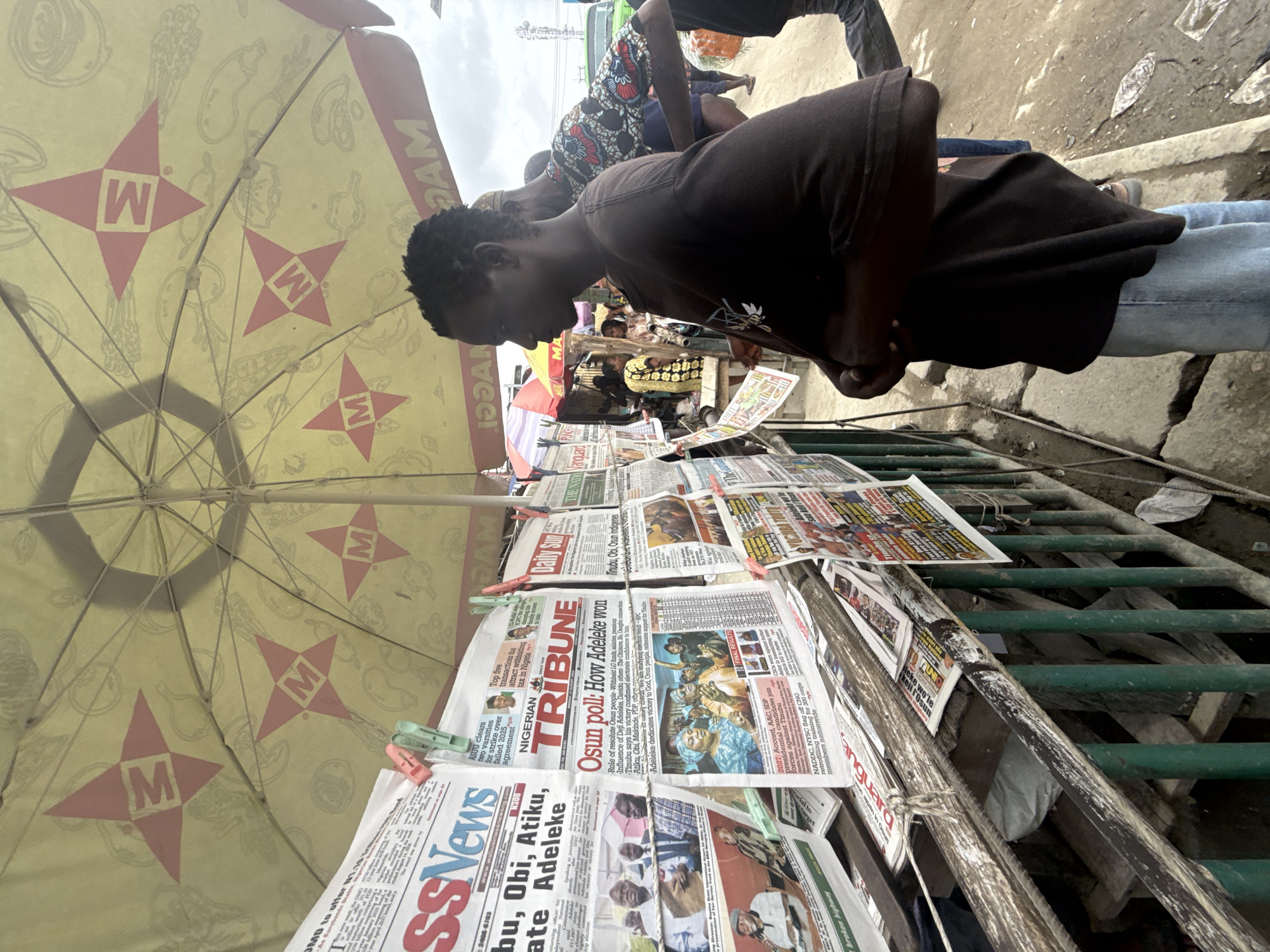 In Lekki, Nigeria, a young Black man stands facing right, wearing a dark t-shirt and light blue jeans, his arms crossed. He is looking at a newspaper stand. The stand displays various Nigerian newspapers, some held up by clothespins. An umbrella with a repeating "M" logo covers the stand. Behind him, other people and stalls are visible, with a green truck in the background. The scene appears to be an outdoor market.