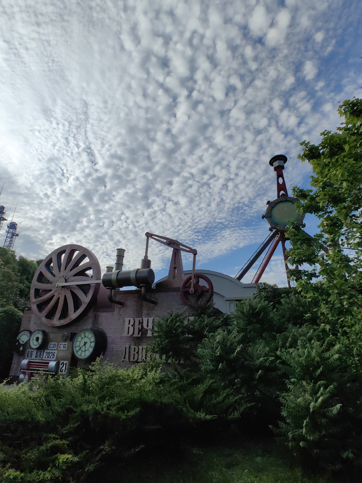 The image depicts a whimsical, steampunk-inspired amusement park attraction in Sirius, Russia. The foreground is dominated by lush green foliage, obscuring the lower portion of the attraction. Rising above the greenery is a large, stylized structure resembling an old-fashioned steam engine, complete with a prominent, oversized wheel on the left and industrial-looking pipes and gauges. The word "ВЕЧ" (VECH) is visible on the side of the structure, which is likely part of a longer word or phrase. To the right of the steam engine structure, a tall, slender amusement ride structure with a circular element at its top and supporting diagonal beams is visible. The background features a bright blue sky partially filled with white, fluffy clouds. In the distance, to the left, are tall, lattice-like tower structures, possibly part of another attraction or infrastructure. The overall scene suggests a recreational area with a unique, imaginative design.
