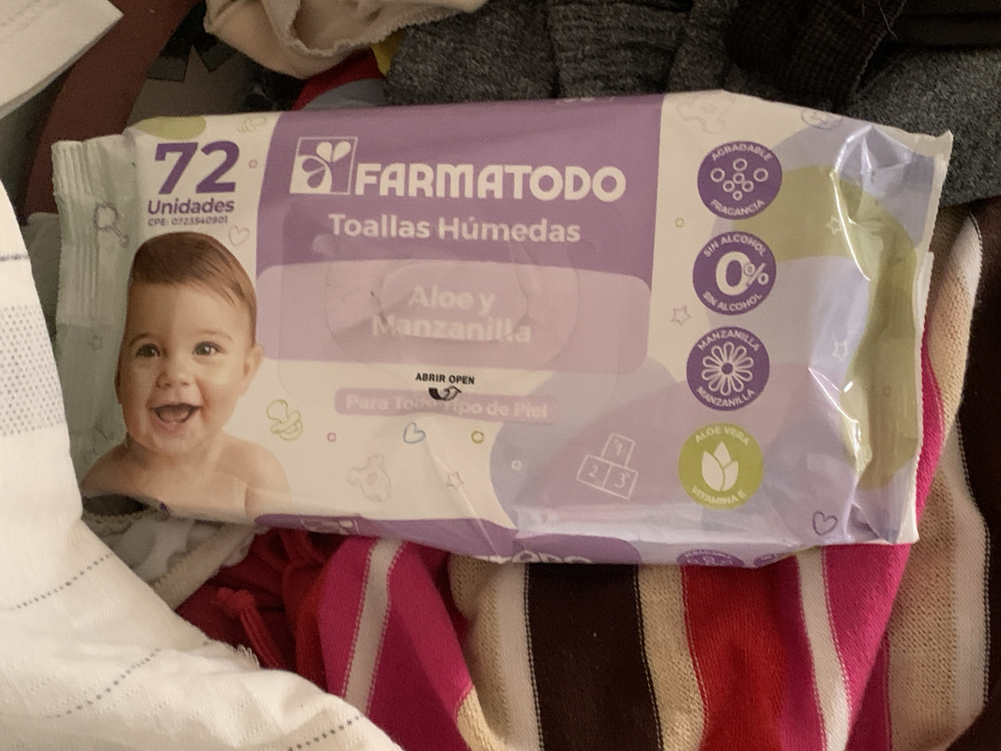 A package of Farmatodo brand Aloe and Chamomile scented baby wet wipes is partially visible, resting on folded striped fabric. The package is predominantly purple and white and features a large image of a smiling baby's face. Text on the package indicates "72 Unidades" (72 Units), "Toallas Húmedas" (Wet Wipes), "Aloe y Manzanilla" (Aloe and Chamomile), and "ABRIR OPEN". Smaller circular icons on the lower portion of the package highlight "SIN ALCOHOL" (Alcohol-Free), "FRAGANCIA AGRADABLE" (Pleasant Fragrance), and "VITAMINA E". The scene is set indoors, with hints of other fabrics and dark surfaces suggesting it may be within a home or storage area. The exact location in Santa Ana, Venezuela, cannot be determined from the provided image.