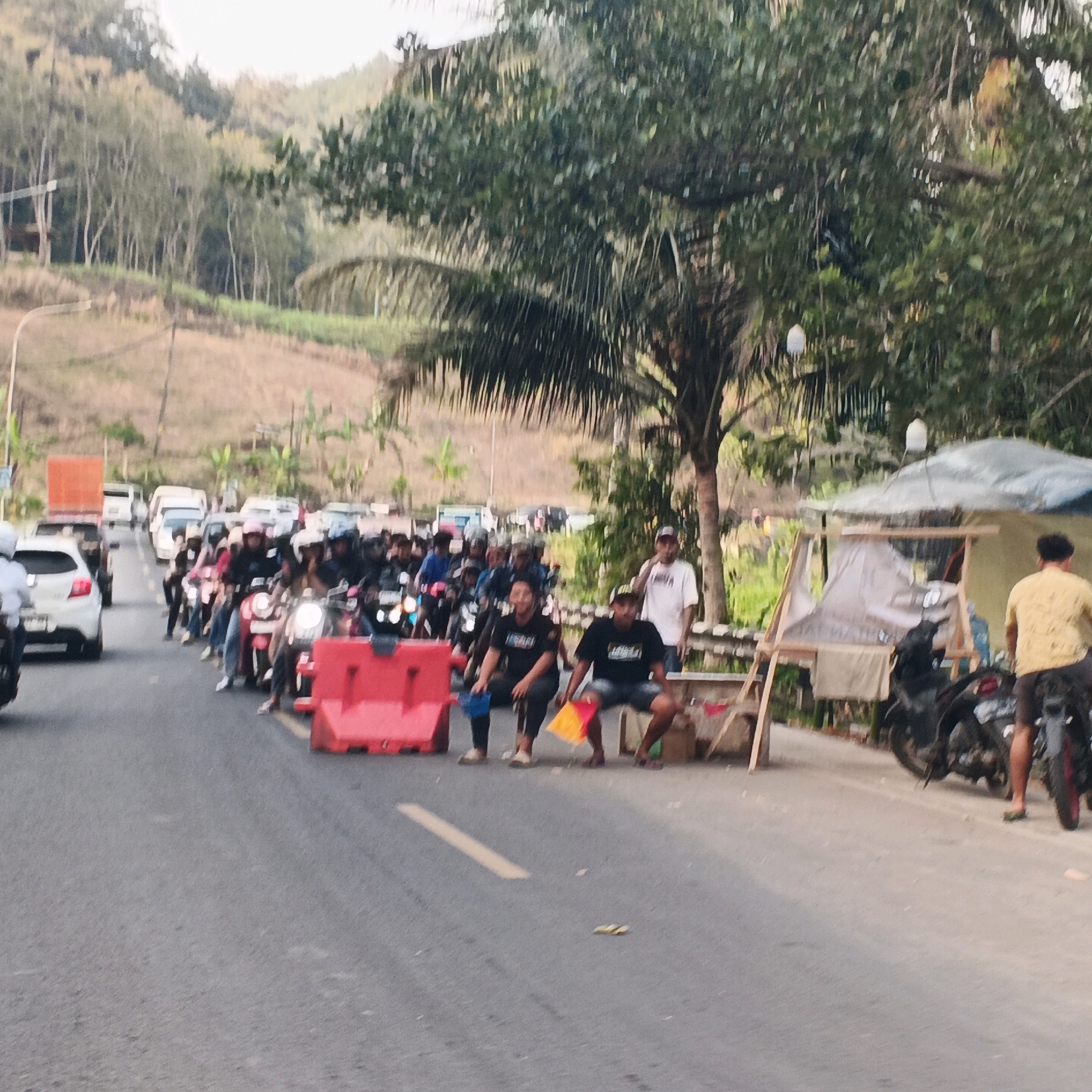 The image depicts a traffic jam on a road in Kebasen, Indonesia. A long line of motorcycles and cars are stopped on the asphalt road, indicating a standstill. Several people are visible, some on motorcycles wearing helmets, and others standing or squatting by the roadside. Two individuals in the foreground are seated, one holding a blue bucket and the other a red and yellow flag, suggesting they might be directing traffic or involved in an event. Red traffic barriers are placed on the road, further emphasizing the congestion. In the background, a densely wooded hillside with scattered trees and vegetation rises. A small, makeshift shelter with a tarpaulin roof stands to the right of the road, and a few more motorcycles are parked nearby. The overall scene suggests a busy and possibly chaotic traffic situation in a semi-rural or developing area.