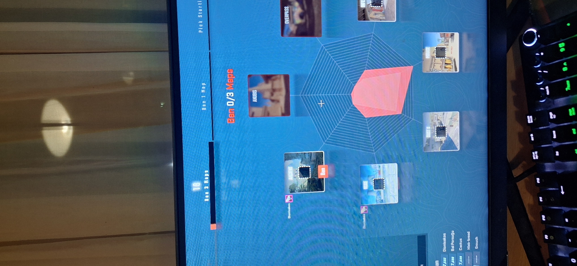 The image shows a monitor displaying a game menu where the user is in the process of banning maps. The menu indicates "Ban 0/3 Maps" and shows a selection of maps, including "Anubis", "Overpass", "Mirage", "Dust II", "Ancient", and another map partly obscured. "Ancient" is selected to be banned. Additional menu elements are visible at the bottom, displaying options and names like "Dionisakos", "Bal Porsuğu", "Cactus", "Hleb-bread", and "Grouch". Part of a keyboard with illuminated keys is visible on the right side of the image. The image does not show Burgas, Bulgaria.