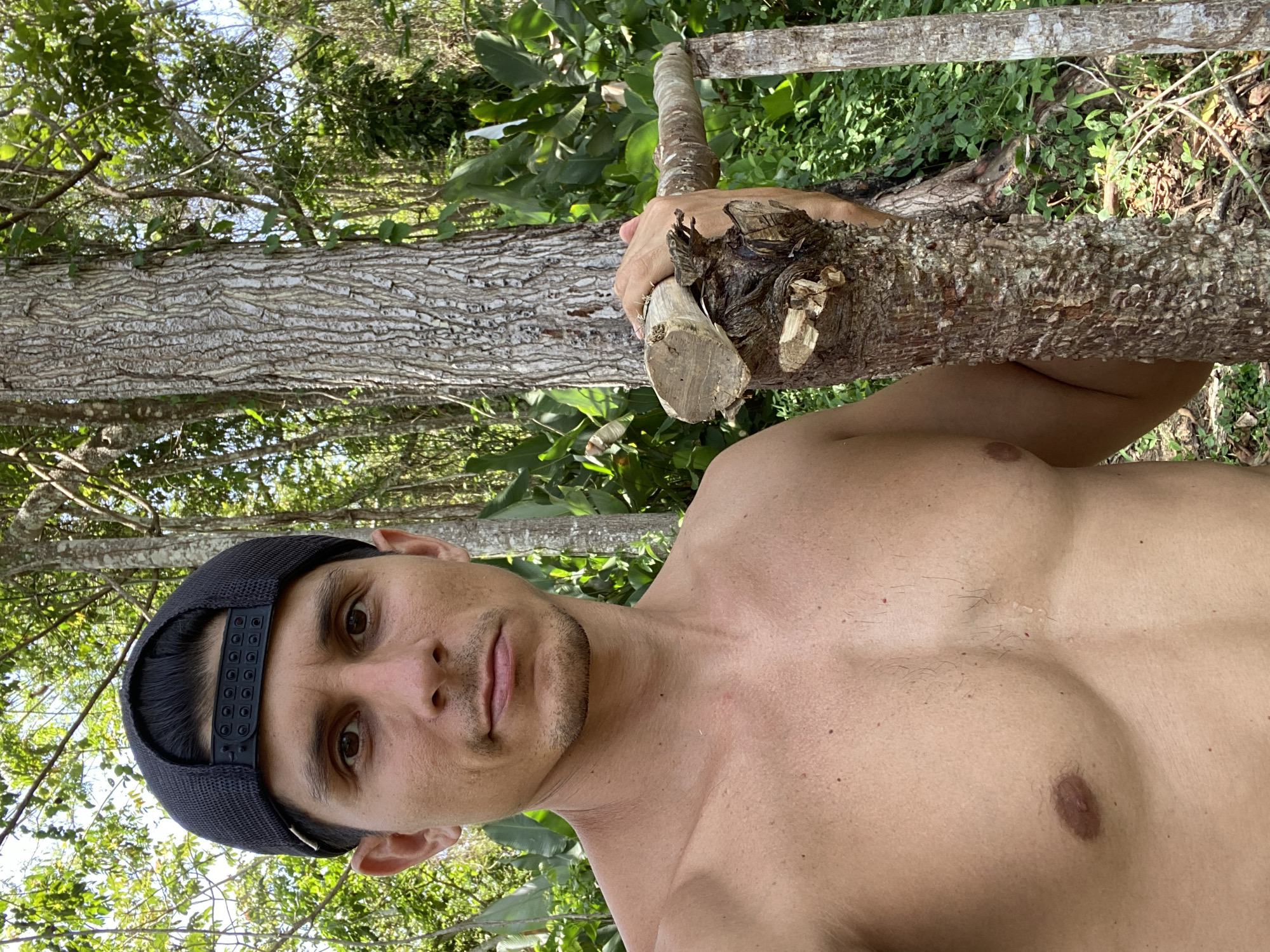 A shirtless man wearing a black baseball cap faces the camera while holding a tree branch. He is outdoors in a wooded area, which suggests he is in the city of Los Velásquez, Venezuela. The man appears to be in his late 20s or early 30s, with brown eyes and short dark hair under the cap. He is holding a medium-sized tree branch, which has a rough texture. The background consists of trees and foliage, which are commonly seen in the city of Los Velásquez, Venezuela.