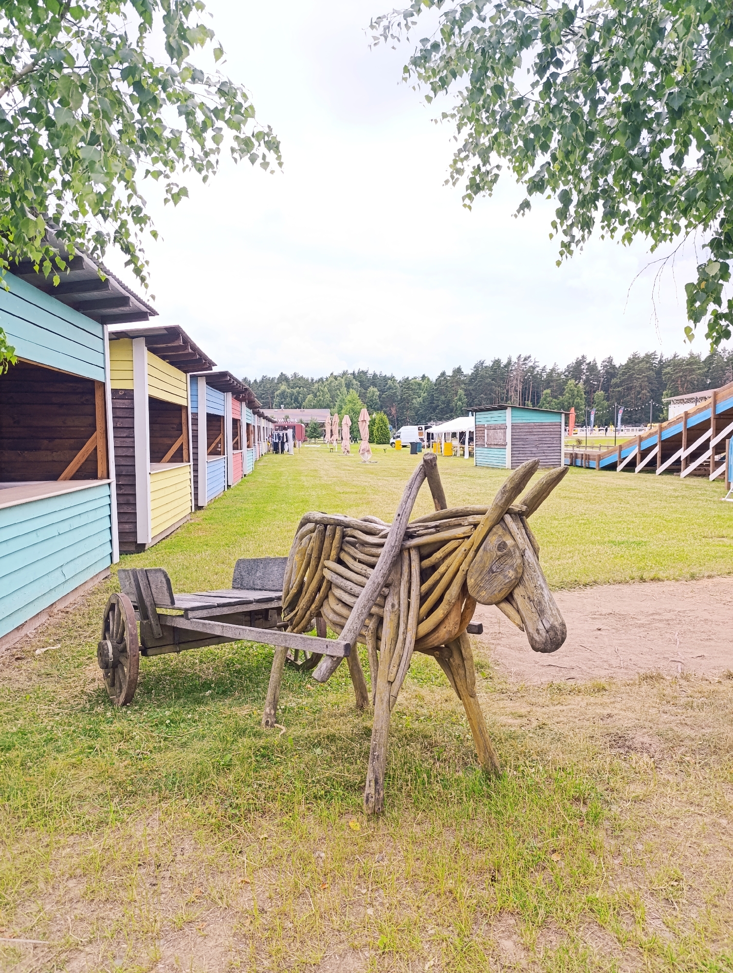 The image depicts an outdoor scene, likely a fairground or festival, with various structures and decorations. In the foreground, a rustic wooden sculpture of a donkey pulling a cart is prominently displayed. The donkey is made from intertwined branches, and the cart is a simple wooden structure with large wooden wheels. The scene is set on a grassy area with a dirt path in front of the sculpture.
Behind the sculpture, a row of colorful, wooden stalls stretches into the background. These stalls are painted in bright hues of blue, yellow, and pink, suggesting a commercial or entertainment venue. Further in the distance, more tents and structures can be seen, along with a forest of pine trees. There are also bleachers or tiered seating visible on the right side of the image.
The sky is overcast, indicating a cloudy or possibly rainy day. The overall atmosphere appears to be that of an outdoor event, with the wooden decorations and stalls contributing to a festive or rustic ambiance. While some people are visible in the distance near the stalls, they are too far away to discern any specific activities or emotions. There is no visible text in the image. The time of day is unclear due to the overcast sky.