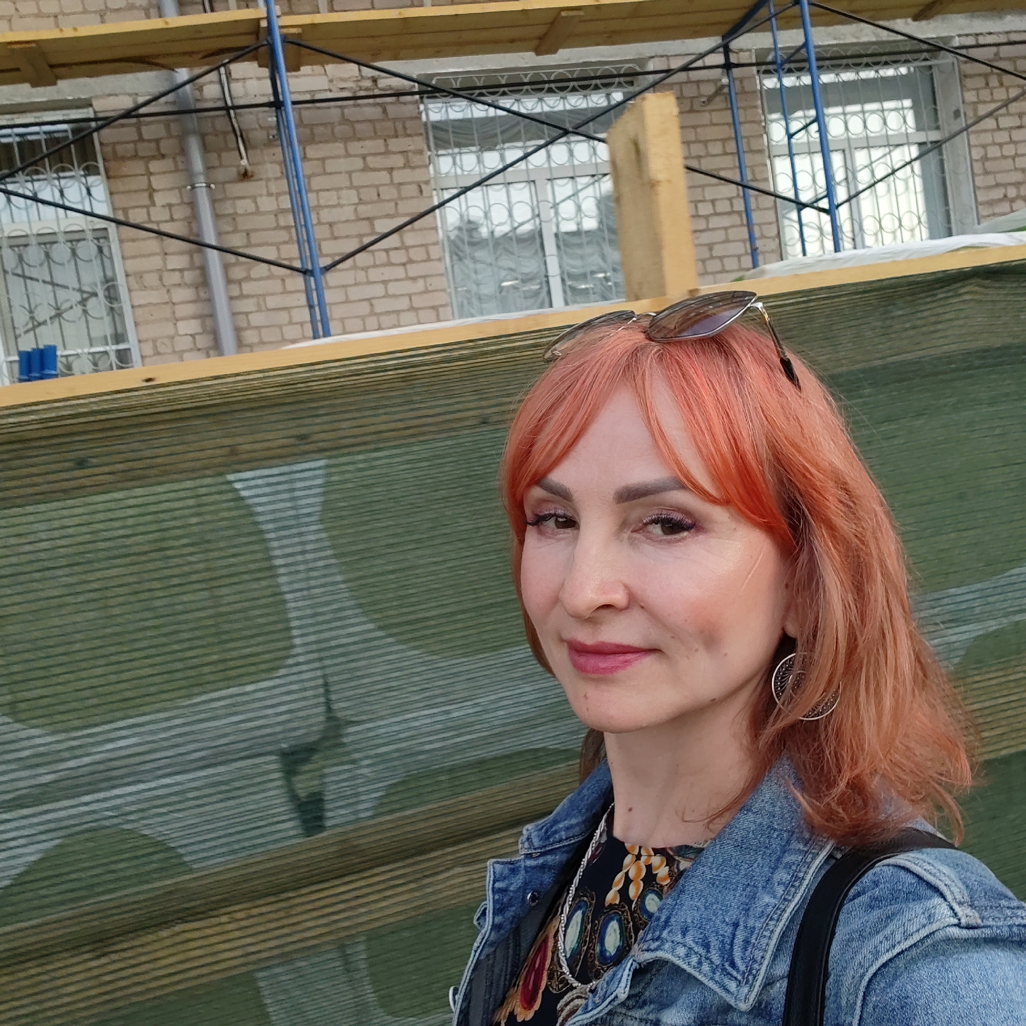 A woman with dyed orange hair and sunglasses perched on her head is centered in a medium shot. She wears a denim jacket over a floral-print dress and large hoop earrings. The background is a construction site with scaffolding against a brick building with barred windows. Green and brown camouflage netting partially obscures the scene. The location is in the city of Chaikovsky, Russia.