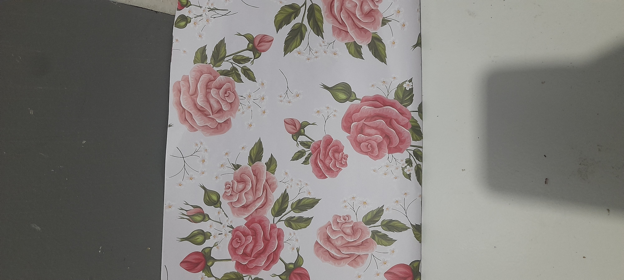 The image shows a section of floral-patterned wallpaper featuring pink roses, green leaves, and small white flowers. The background is light-colored, possibly white or a very pale shade. The wallpaper is positioned between a gray surface on the left and a white surface on the right. The location and city of Buketteukuh, Indonesia are not evident within the image. There are no people, subjects, actions, or interactions visible.
