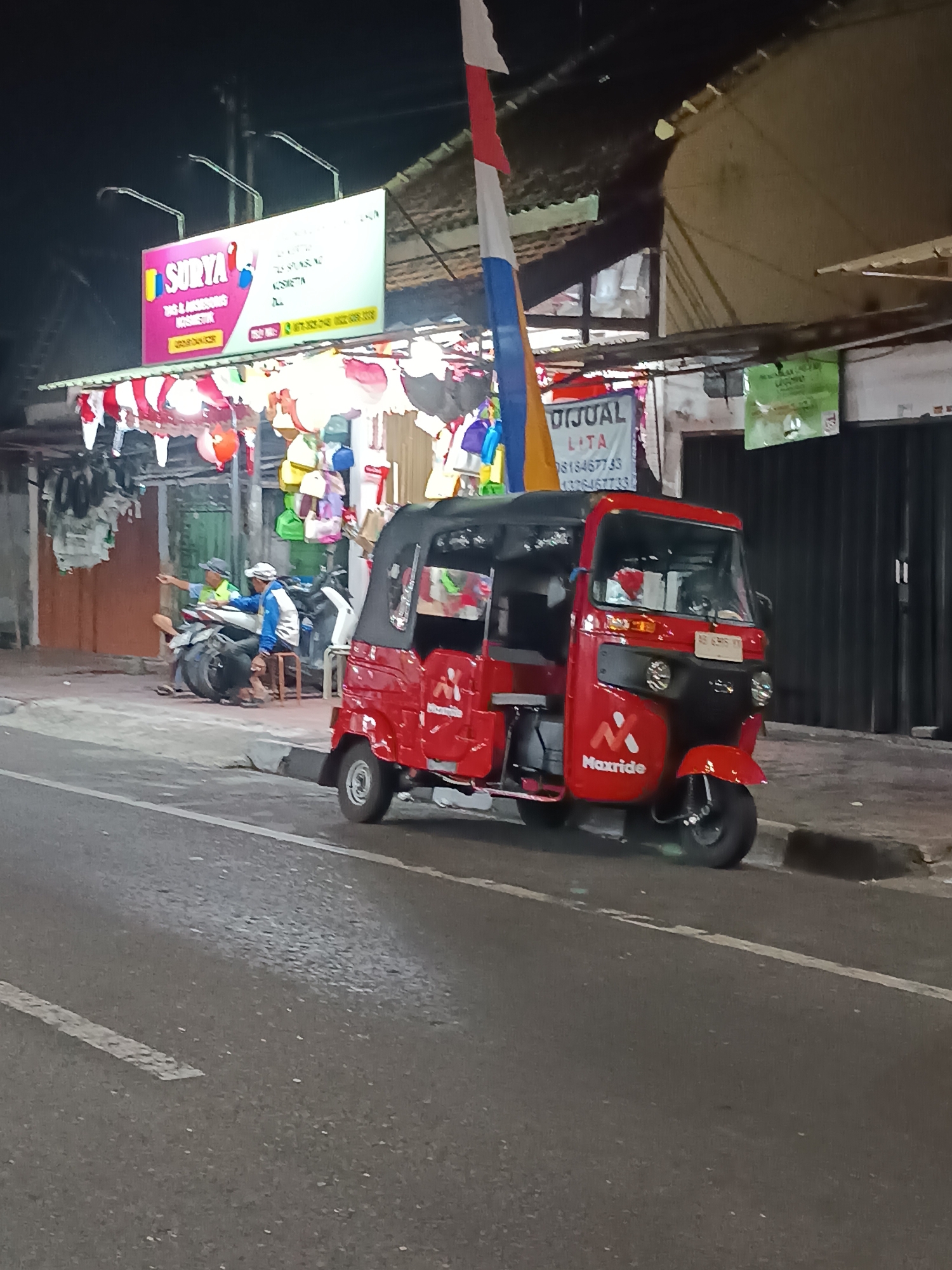 A red three-wheeled vehicle, identified as a Maxride, is parked on the side of a street at night. The vehicle is positioned slightly angled towards the viewer, with its front wheel clearly visible. In the background, a storefront is illuminated by artificial lighting, displaying various goods, including what appear to be balloons and bags. A sign above the storefront reads "SURYA" in large pink letters, and below it, "TAS & AKSESORIS KOSMETIK" (Bags & Cosmetic Accessories). Another sign in Indonesian reads "DIJUAL" (For Sale). Two men are seated on a motorcycle near the storefront. The scene appears to be in a commercial area of Yogyakarta, Indonesia, given the Indonesian text on the signs. The street is paved, and a white line marks the edge of the roadway.