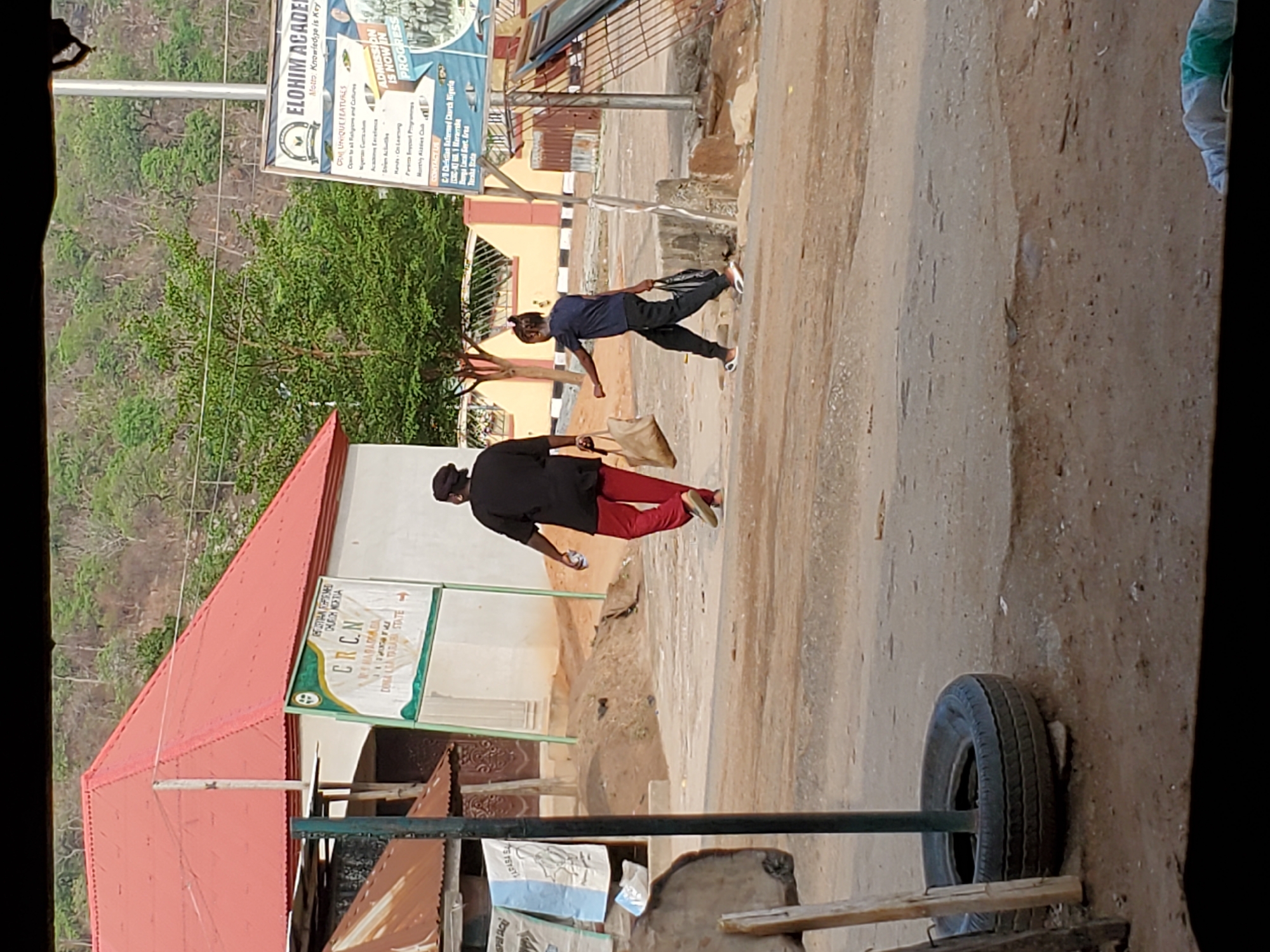 Two people walk along a dusty street next to a building with a red roof and a sign for C.R.C.N church. A sign for Elohim Academy is visible on a pole, indicating the location is Mararaba, Nigeria. Debris and an old tire sit in the foreground. One person wears a black shirt and red pants, while the other wears a dark shirt and pants.
