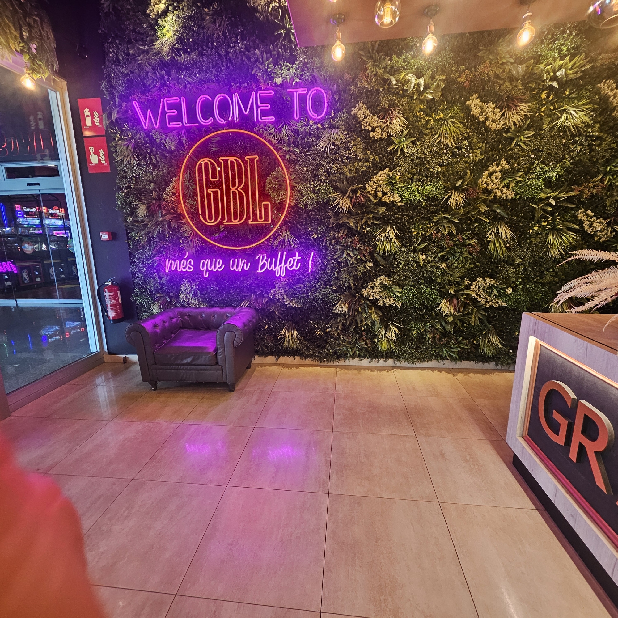 This image captures the interior of a commercial establishment located in Salt, Spain, likely an entrance or waiting area. The dominant feature is a large artificial vertical garden wall, composed of various shades of green and some lighter-toned foliage, covering the central and right sections of the back wall. Mounted on this plant wall are three neon signs: "WELCOME TO" in purple capital letters at the top, a circular orange-red neon sign with the letters "GBL" centrally positioned, and "més que un Buffet !" in purple cursive script below.
To the left of the plant wall, on a dark gray vertical surface, two red rectangular safety signs with white pictograms (a fire extinguisher and a fire alarm pull station) are affixed, with a red fire extinguisher mounted beneath them. Adjacent to this wall is a glass door or window, reflecting a brightly lit interior space with multiple gaming machines or arcade elements visible through the reflections.
A dark, tufted Chesterfield-style armchair is positioned on the light-colored tiled floor beneath the neon signs. In the bottom right foreground, a counter with a wooden top and a dark front panel displaying the orange letters "GR" is partially visible. The ceiling above features three exposed-bulb pendant lights emitting a warm glow. The tiled floor, light beige or tan, reflects the purple and orange light from the neon signs. No individuals are present in the scene; however, a blurred red/pink object is visible in the bottom-left corner of the frame, possibly part of the photographer's hand or clothing.