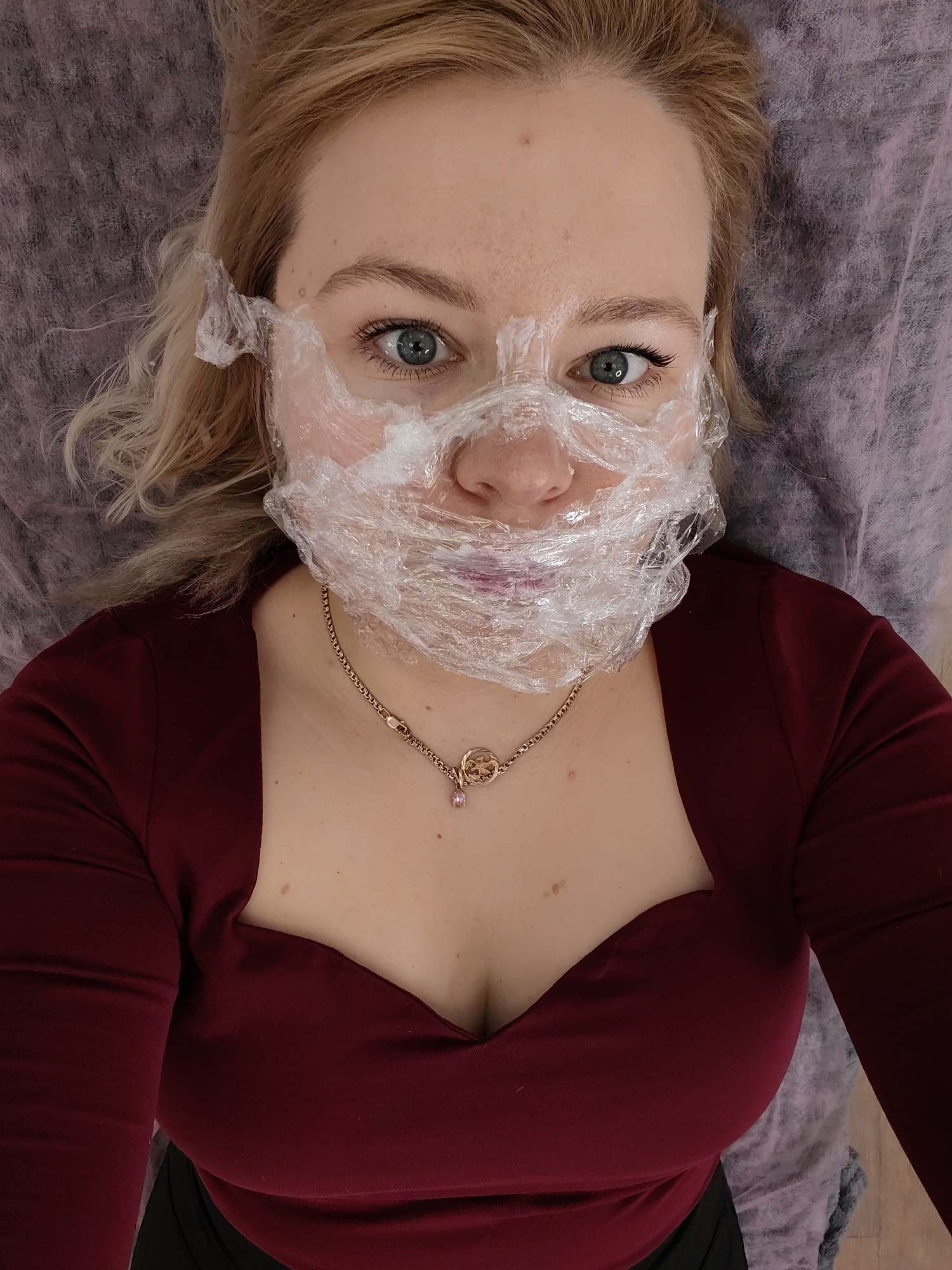 A fair-skinned woman with blonde hair and striking blue-green eyes lies on her back, gazing directly into the camera. Her face is mostly covered with clear plastic wrap, a common practice during beauty or medical treatments like facials or numbing cream application, which leaves her eyes and part of her mouth visible. Small, subtle blemishes are visible on her forehead, and her eyelashes appear long and defined.
She is dressed in a dark red, long-sleeved top featuring a low, sweetheart neckline. A delicate gold chain adorns her neck, from which hangs a circular pendant with a small pink gem. Her lips have a reddish tint. She is positioned against a background of a light purple, textured disposable sheet or covering, typical of a salon, spa, or clinic setting. Her expression is direct and alert. No text is visible in the image, and the indoor setting does not provide cues for the time of day or weather. This scene takes place in Izhevsk, Russia.