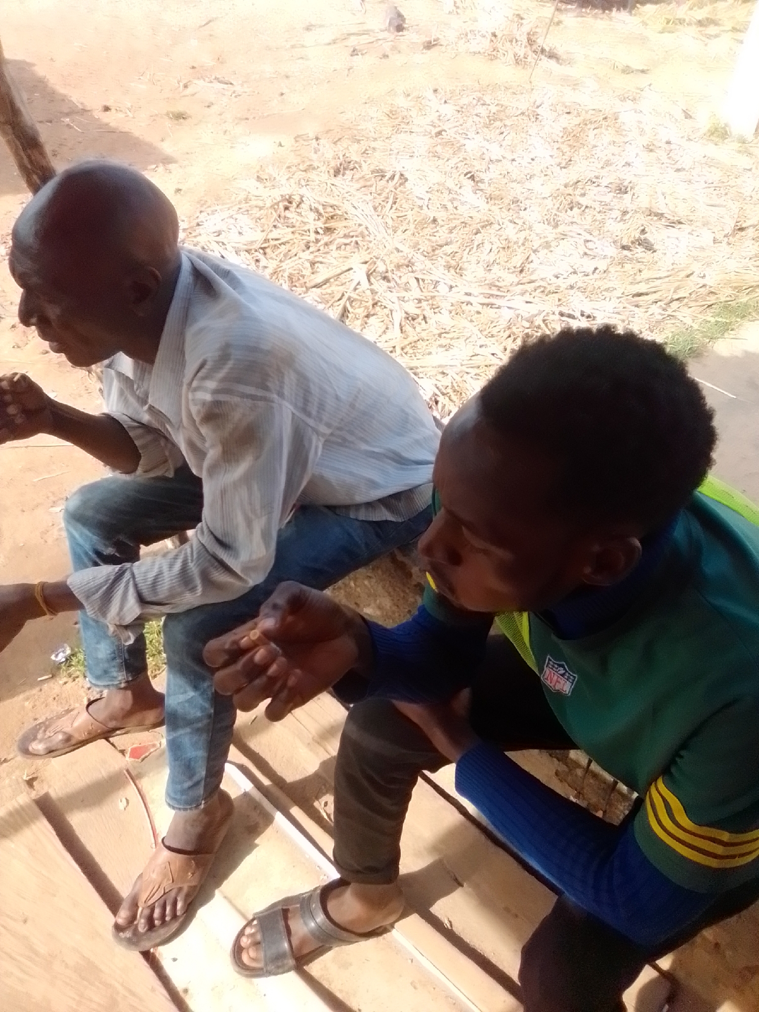 In Pantame, Nigeria, two individuals are seated outdoors on a wooden surface, which appears to be part of a raised platform or bench. The ground is covered with dry vegetation and sand.
The person on the left is an older man with a bald head and dark skin, wearing a light blue and white striped long-sleeved shirt and blue jeans. He is also wearing brown flip-flops. His hands are clasped together in his lap, and he appears to be looking down or to the side.
The person on the right is a younger individual with dark skin and short, dark hair, wearing a green long-sleeved shirt with blue sleeves and yellow stripes. The shirt has an NFL logo patch on the chest. This person is wearing dark pants and black sandals. Their hands are visible, with the right hand extended slightly forward, fingers open, and the left hand appearing to be near the right hand. They are looking towards their hands.
The lighting suggests it is daytime, with sunlight casting shadows. The overall scene depicts a casual outdoor setting in a dusty environment.