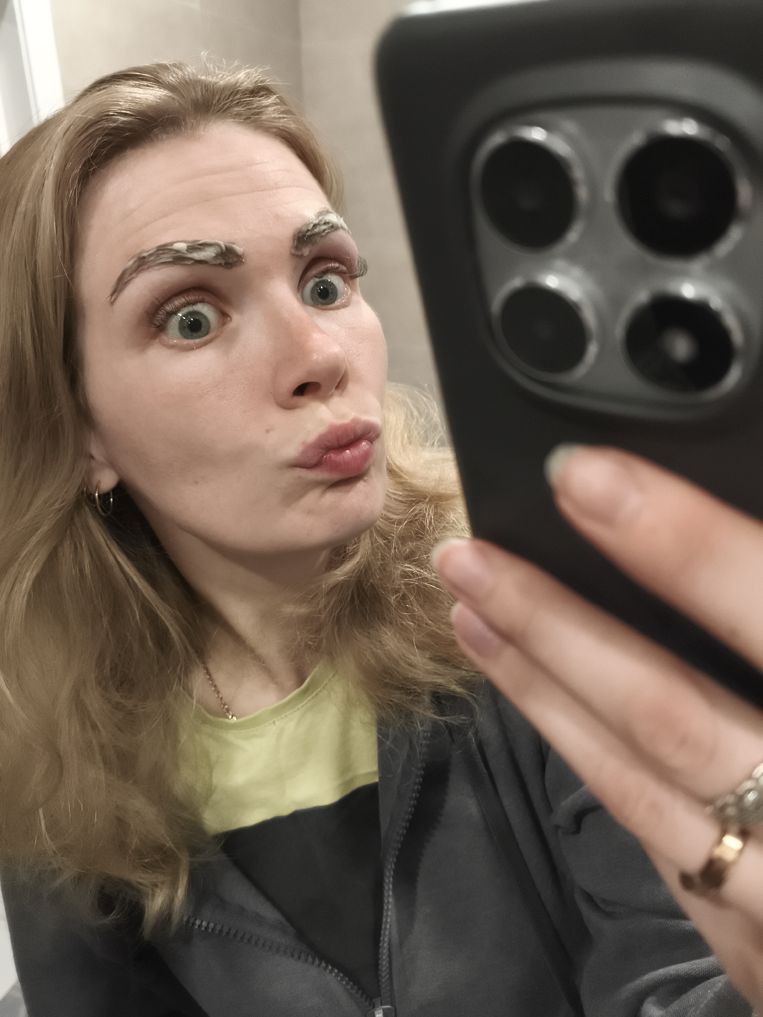 The image depicts a woman taking a selfie indoors, potentially in a bathroom given the tiled wall. She has fair skin, blonde hair, and green eyes. Her eyebrows are covered in a light-colored cream or paste. She is making a puckered-lip expression. She is wearing a light green and dark gray shirt or jacket. She is holding a black smartphone with multiple rear cameras. Her left hand has a gold and a silver ring on her fingers. A small gold hoop earring is visible in her left ear. A thin gold chain necklace is around her neck. The location is not identifiable as Русский Вожой, Russia, from the image alone.