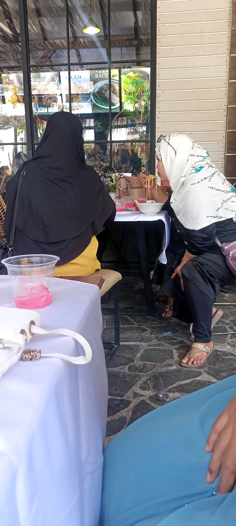 In Desa Ponokawan, Indonesia, two women are seated at a table. The woman on the left wears a black hijab and a yellow top, facing away from the camera. The woman on the right, wearing a white hijab with blue accents, is actively eating noodles with chopsticks, leaning over a white bowl. A pink drink is visible in a plastic cup on a white tablecloth in the foreground, and a white handbag rests on the same tablecloth. A glass table with a dark base is situated between the women. The background shows a window with a reflection of signage and the interior of a building. The ground is paved with dark, irregular stones.