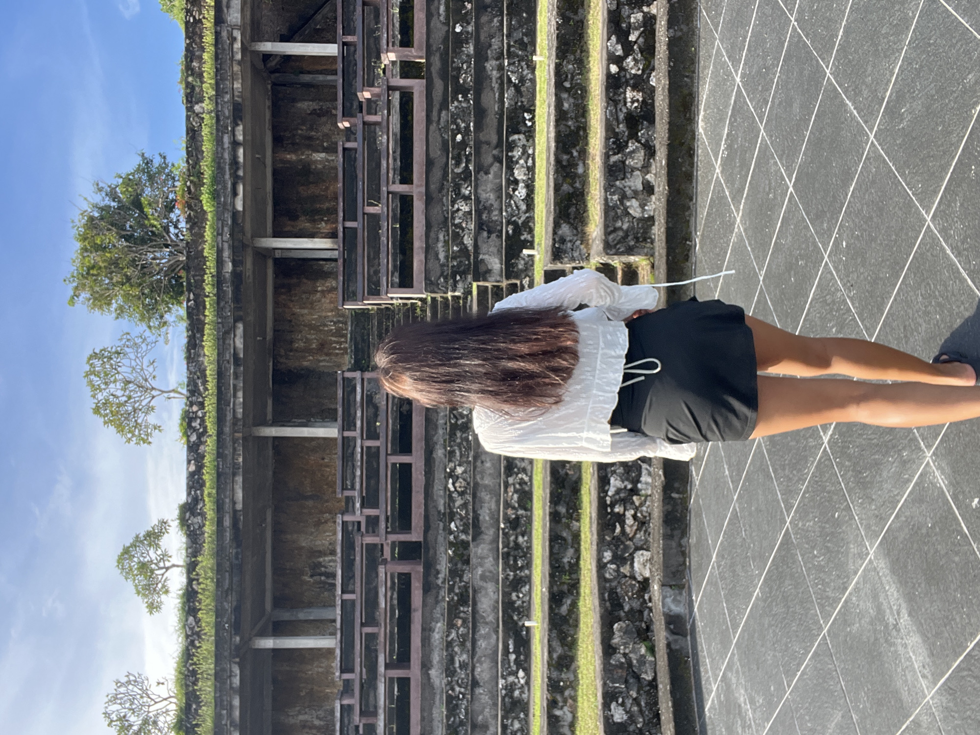 The image captures a person from behind, standing outdoors in what appears to be a historical or temple complex. The individual has long, dark brown hair and is wearing a white long-sleeved top and a black skirt or shorts.
The scene features a large, multi-tiered structure in the background, resembling ancient steps or terraces made of dark stone or concrete. These tiers are partially covered with green moss or small plants, and some sections show exposed wooden or metal beams. Further back, a wall or raised platform is visible, topped with lush green vegetation. Several trees with light-colored trunks and green foliage stand out against a clear blue sky, suggesting a tropical environment; some appear to be frangipani trees.
The ground where the person is standing is paved with dark grey square tiles, laid in a diamond pattern highlighted by lighter grout lines. The bright, clear blue sky indicates it is daytime, likely a sunny morning or afternoon. The person is facing away from the camera, seemingly observing the ancient structures and natural surroundings. There is no discernible specific activity other than standing and viewing the site. The image evokes a sense of exploration or tourism in a historical and natural setting. No text is visible in the image.