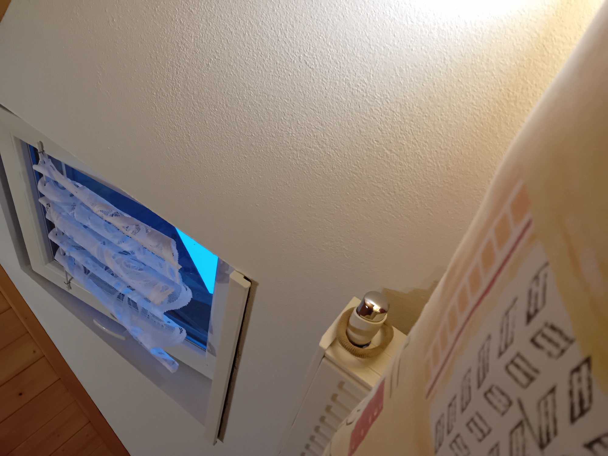 The image captures an interior view of a room, likely an attic due to the sloped wooden ceiling visible in the upper left corner. A dormer window with white trim is open, allowing bright blue light to enter, suggesting it is daytime. White lace curtains are drawn across the window, adding a delicate texture to the scene.
In the foreground, a rolled-up object, possibly a blind or a large print, is partially visible. It has a cream-colored background with a repeating pattern of dark rectangular shapes and thinner red and orange horizontal lines. Below this, a white radiator with visible fins runs horizontally. Mounted on the radiator is a small, silver, cylindrical object with a chrome knob, which could be a thermostat control.
The walls appear to be textured and painted a light color. The lighting in the room seems to be a mix of natural light from the window and artificial light, possibly from a lamp, casting a warm glow on the wall to the right. The perspective is from a low angle, looking upwards towards the window and ceiling. The location is identified as Zuberec, Slovakia.