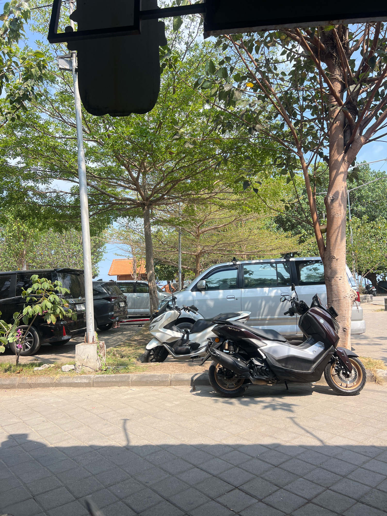 The image depicts a parking area with several vehicles, including two motorcycles and three cars, under the shade of trees. In the foreground, two scooters are parked side-by-side on a paved surface. The scooter on the right is dark maroon and silver, while the one on the left is predominantly white. Behind the scooters, a silver minivan is parked, with its windows reflecting the surroundings. To the left of the minivan, two black SUVs are parked. The environment suggests a sunny day, with shadows cast by the trees and vehicles. In the background, glimpses of buildings with orange roofs and the sky are visible. The scene is consistent with a parking lot in Kuta, Indonesia, a popular tourist destination known for its beaches and vibrant atmosphere.
