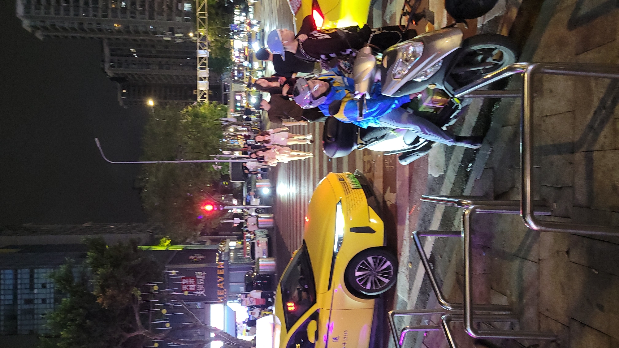 At night in an unidentified urban location, the scene depicts a stationary yellow taxi obstructing a pedestrian crosswalk. The street is illuminated with lights from buildings and traffic signals. A group of people are standing on the crosswalk next to a parked silver scooter with two people on it. A row of silver metal bike racks borders the crosswalk.