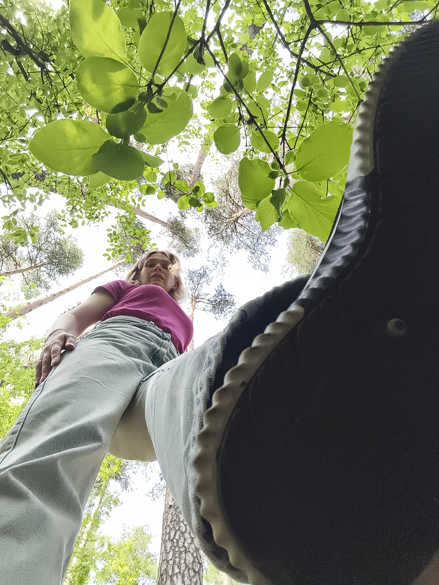 The image is taken from a low-angle perspective, looking up through trees and foliage towards a person. The person is wearing a pink shirt and light-colored pants. Part of a shoe is visible in the foreground, obscuring the view. The scene appears to be in a wooded area with trees visible behind the person. The location in the city of Izhevsk, Russia is not identifiable based on the image content.