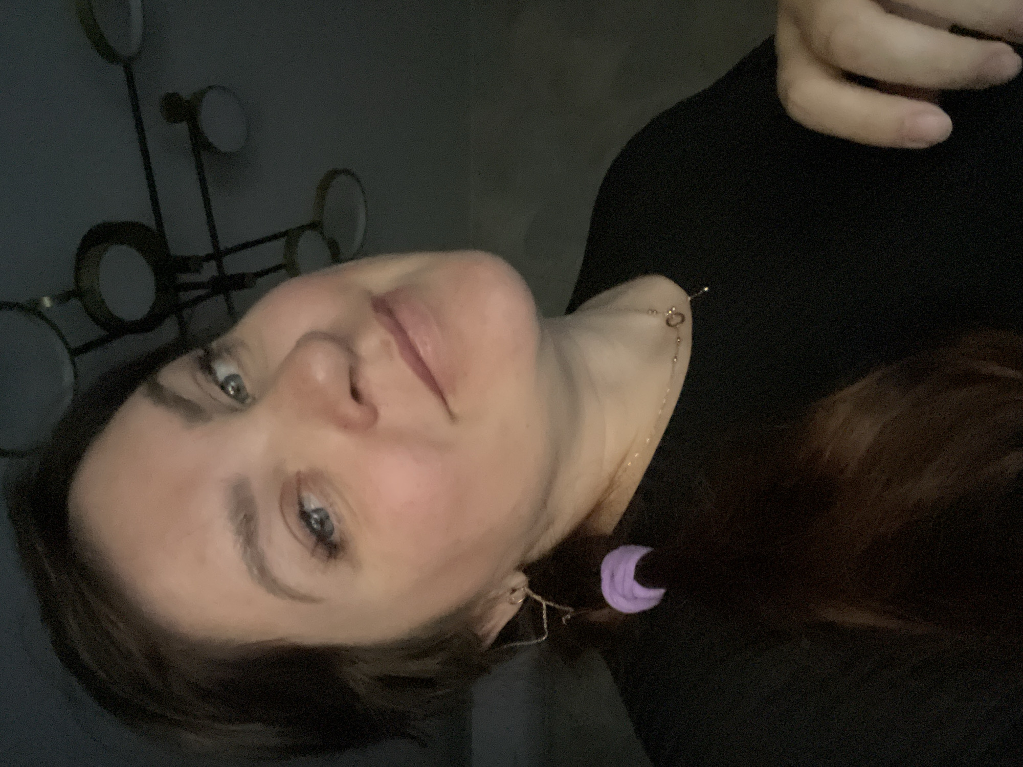 The image is a close-up portrait of a woman, presented on its side, showing her head and upper torso. The woman has fair skin, blue-grey eyes, and dark brown hair. She looks towards the camera with a subtle, pleasant expression and a slight smile. Her face is softly illuminated, suggesting dim, artificial indoor lighting.
She wears a delicate gold-colored chain necklace with a small heart-shaped charm. On her visible ear (on the left side of the rotated image), she has a distinctive earring: a long, thin, gold-colored chain ending in a small, light purple, crescent moon charm. She is dressed in a dark, possibly black, top or sweater. In the top right corner, a hand with fingers is partially visible.
The background features a dark wall adorned with a decorative fixture or light source, composed of multiple circular elements connected by thin rods. These circles appear to have lighter, possibly reflective, centers. The overall scene suggests an indoor setting with an intimate, softly lit atmosphere. There is no visible text in the image.