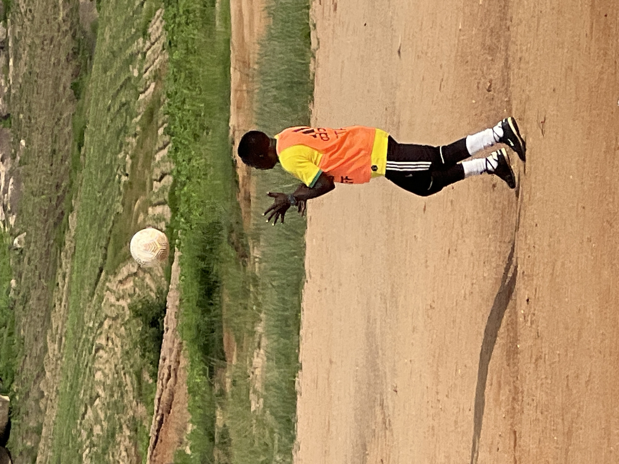 In Jos, Nigeria, a young person is shown in mid-action, playing with a soccer ball outdoors. The individual, wearing a bright yellow jersey with an orange vest over it and black pants with white stripes, is looking upwards and has their hands outstretched, seemingly in the process of catching or heading the ball which is suspended in the air above them. The scene takes place on a sandy, dirt surface, indicative of a makeshift field. In the background, there are terraced, green hills and rocky outcrops, typical of the natural landscape found in parts of Jos. The lighting suggests it is daytime, possibly late afternoon, given the elongated shadow cast by the person on the ground. The overall environment is rural or semi-rural, away from urban development.