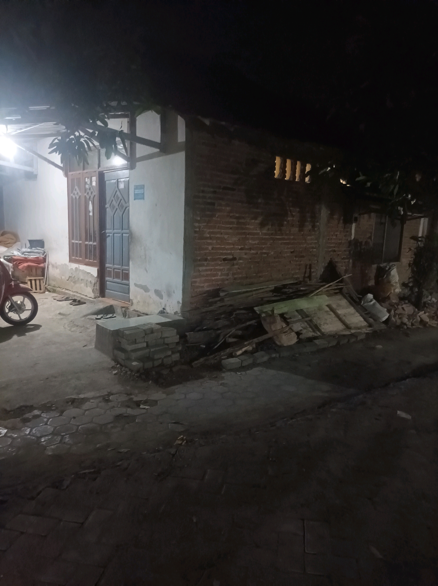 The image depicts an outdoor scene at night, likely in the city of Canga'an, Indonesia. The foreground shows a paved area with hexagonal paving stones, some of which are wet or reflective. To the left, a red motorcycle is parked. Adjacent to the motorcycle is a low-lying structure with a wooden crate containing rolled-up fabric or bedding.
The main structure in the image is a building with a white-painted wall and a brick wall extension. There is a doorway with a double door, featuring decorative patterns. To the left of the door is a window with similar decorative elements. The brick wall section has a series of horizontal openings, possibly vents, and appears to be under construction or in a state of disrepair. Piled in front of the brick wall are various construction materials, including wooden planks, metal sheeting, and what appears to be a large piece of flattened cardboard. Some loose bricks are stacked near the doorway. Trees with dark foliage overhang the scene. The lighting is dim, suggesting it is nighttime, with artificial lights illuminating parts of the building and the motorcycle.
