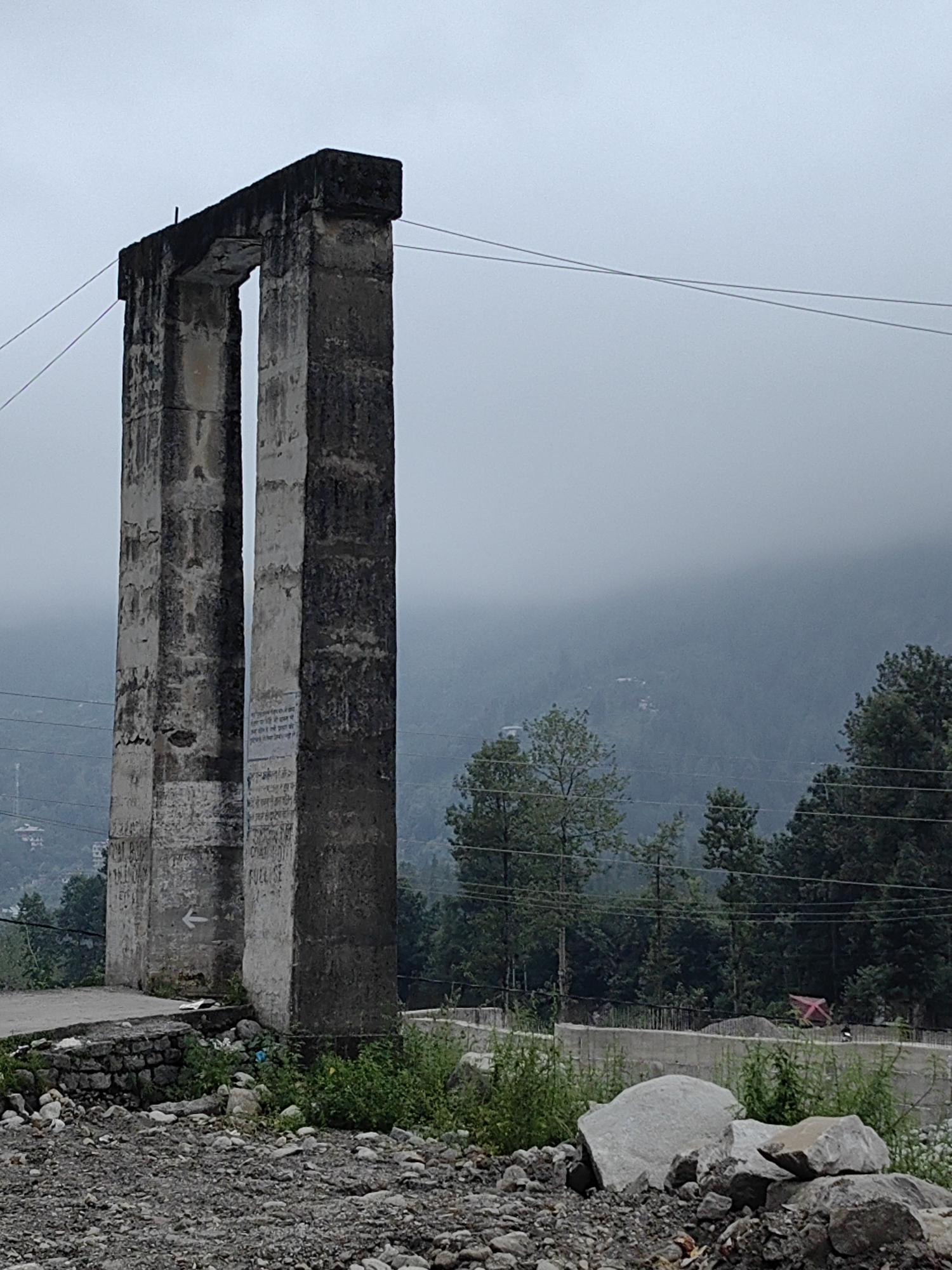An overcast, hazy day in Barheta, India, frames a weathered concrete archway. The archway, composed of two thick, rectangular pillars supporting a horizontal beam, stands at the edge of a paved surface. Faint, unreadable text is visible on the pillars, along with a white arrow pointing left on the left pillar. The ground in front of the archway is covered in small stones and debris. Beyond the archway, a dense forest blankets the undulating hills, their peaks shrouded in mist. Power lines crisscross the scene.