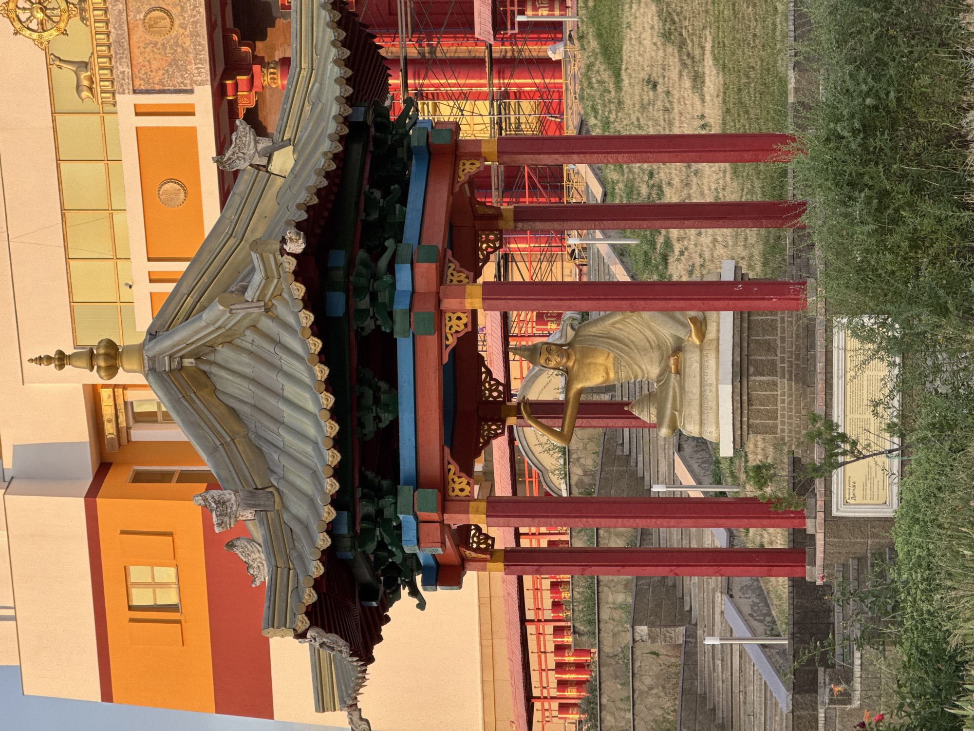 The image depicts a Buddhist temple complex in Elista, Russia. In the foreground, a golden statue of a seated figure, likely a religious icon, is housed within a red and gold pagoda with a multi-tiered, green-tiled roof. The pagoda features ornate carvings and decorative elements. Behind the pagoda, a large, modern building with orange, red, and white striped walls is visible. This building has several windows and a golden spire at the top. The ground is covered in grass and some stone steps lead up to the temple. In the background, scaffolding is apparent on parts of the building, suggesting ongoing construction or renovation. A plaque with Cyrillic script is visible at the base of the pagoda. The overall scene is bathed in warm, late afternoon sunlight.