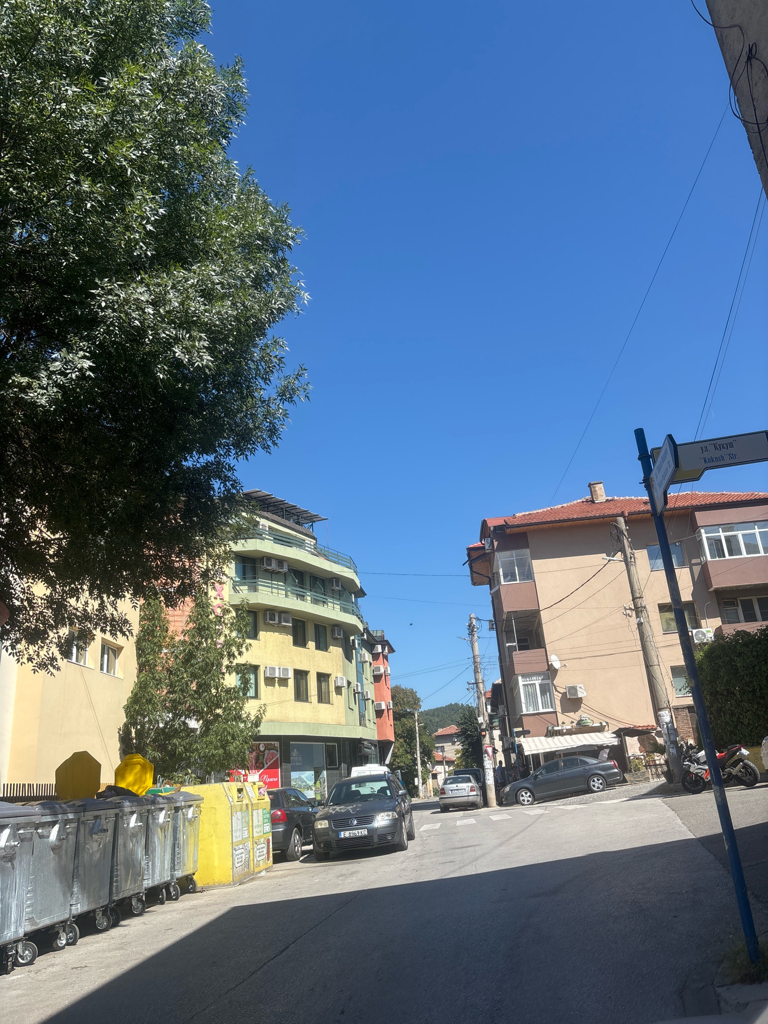 The image depicts a street scene in Blagoevgrad, Bulgaria, on a clear, sunny day. The street slopes downwards from right to left. A large tree with lush green foliage dominates the left side of the frame, casting a shadow across the street.
In the foreground on the left, a row of large, wheeled trash bins is lined up along the sidewalk. These bins appear to be made of metal and plastic, with one prominent yellow bin among them.
Several buildings line the street. On the left, a multi-story building with a light yellow facade features multiple windows and air conditioning units. Adjacent to it, another building exhibits a curved facade with a light green trim and numerous balconies, also equipped with air conditioners.
On the right side of the street, a multi-story building with a light brown and darker brown facade is visible. A street signpost stands in front of this building, displaying text in both Cyrillic and Latin alphabets, indicating "ул. 'Кукуш'" and "Kukush Str.". Further down the street, other buildings and a hillside with trees are visible.
Several vehicles are parked or driving on the street. A dark colored sedan with a license plate starting with "E 8969" is parked on the right side of the street. Another vehicle is visible further down the street, and a motorcycle is parked near the sidewalk on the right. The road surface is asphalt. The sky is a vibrant, cloudless blue. There are power lines and utility poles present in the scene. No people are clearly identifiable.