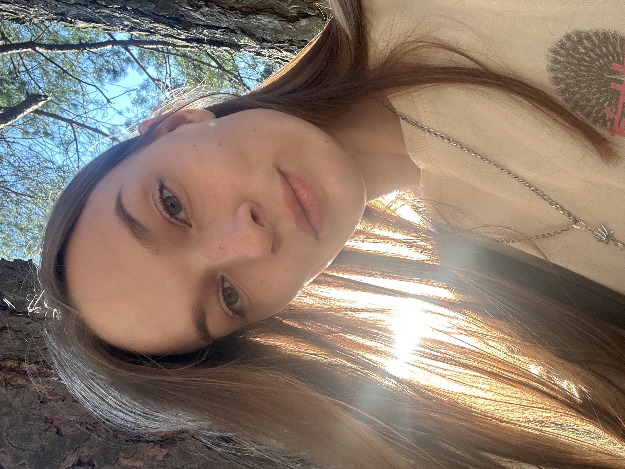 The image is a selfie taken outdoors, with the subject's face tilted upwards and to the side. The person appears to be a young female with long, brown hair, wearing a light-colored shirt and a silver necklace. Sunlight is streaming from the upper left, creating lens flare and highlighting strands of her hair. Behind the subject, tree bark is visible on the left, and a bright blue sky with some tree branches can be seen in the upper portion of the frame. The overall impression is a casual, candid moment captured in natural light. The time of day appears to be daytime, likely late morning or afternoon, given the angle and intensity of the sunlight. There is no visible text in the image.