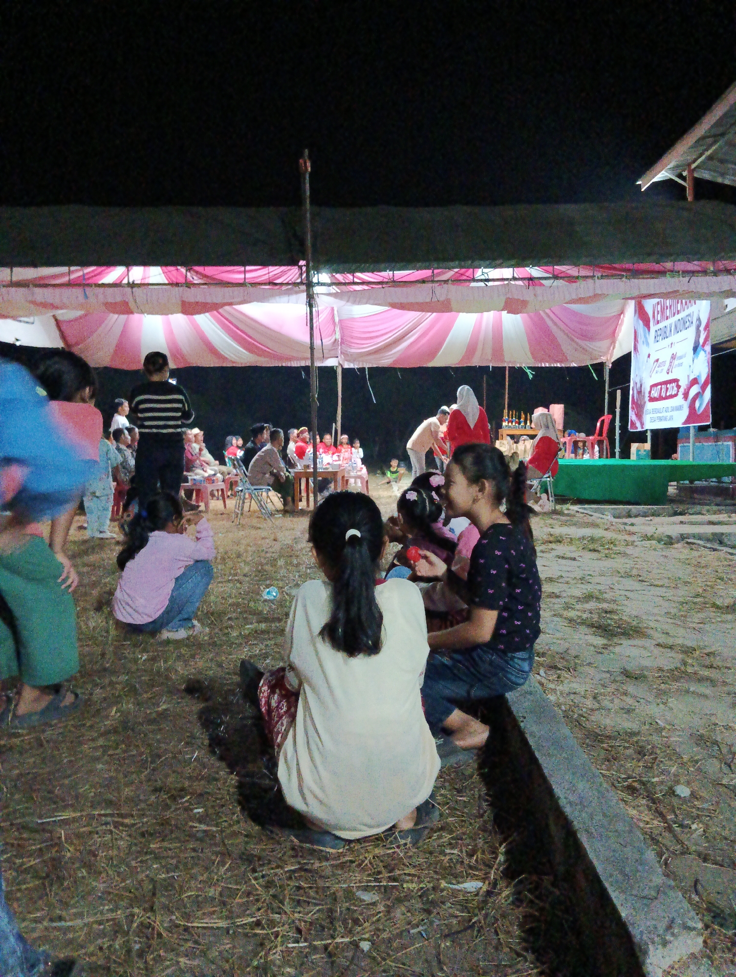 The image depicts an outdoor gathering at night, likely a community event in Pematang Jaya, Indonesia, as indicated by a banner in the background that reads "KEMERDEKAAN REPUBLIK INDONESIA HUT RI 2016 DESA PEMATANG JAYA." A pink and white striped tent covers a stage area where several adults are present, some appearing to be seated at a table and others standing. A banner celebrating Indonesia's Independence Day is prominently displayed on the right. In the foreground and midground, a group of children are seated on the ground, some interacting with each other. One girl in a light-colored shirt and patterned skirt is facing away from the camera, while another girl in a black shirt with a floral pattern and jeans is facing the camera and smiling. A girl in a pink shirt and blue jeans is crouched on the ground. The ground is covered with dry grass and dirt. To the right, a concrete ledge serves as seating for some of the children. The overall atmosphere appears to be festive and communal.