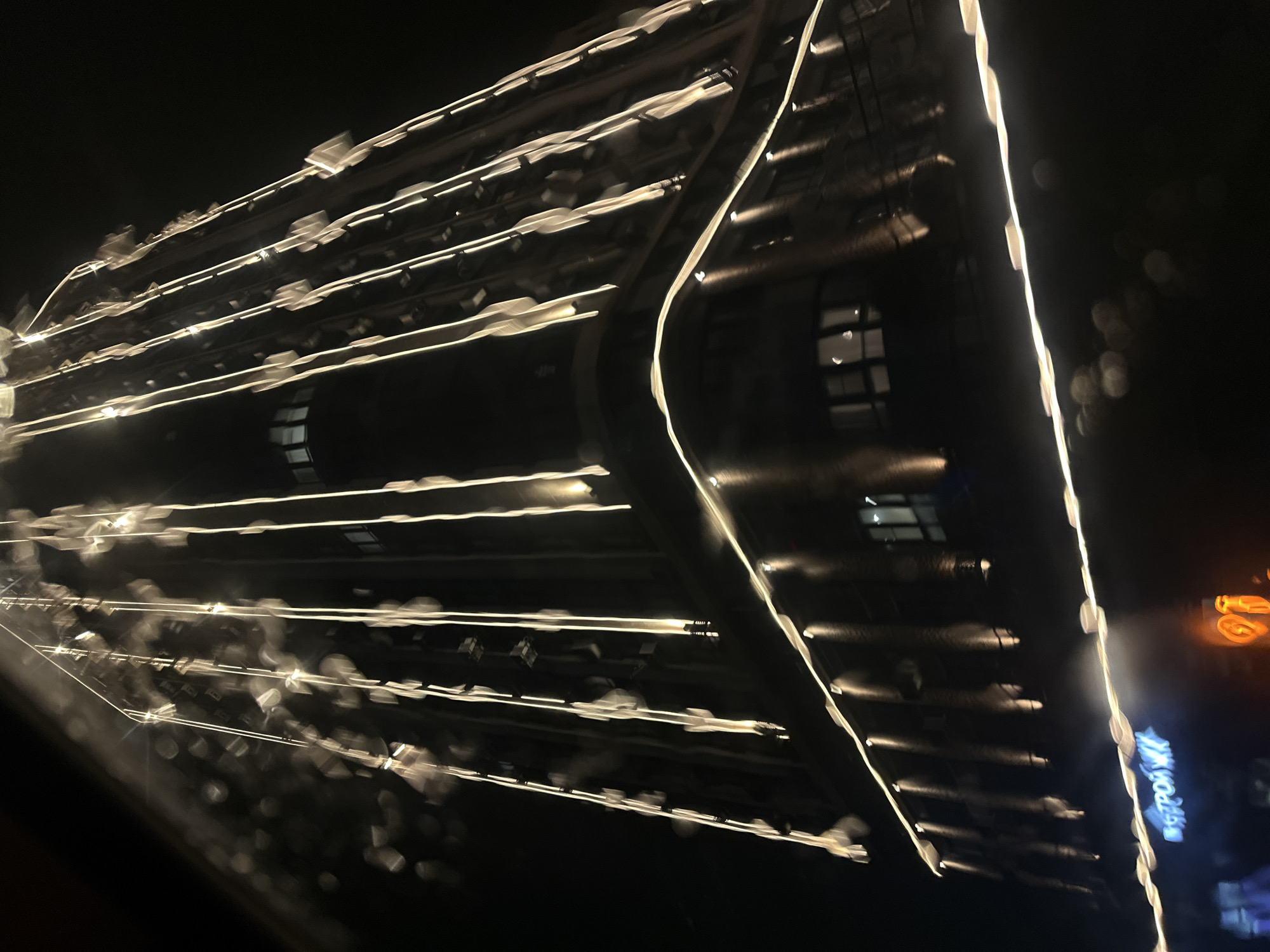 The image shows a night scene, likely taken from a moving vehicle, as evidenced by the light streaks. The subject is a building illuminated with linear lights along its façade. The architecture suggests a modern, possibly commercial, structure. There are windows visible, hinting at multiple floors. The blurred lights on the right suggest other signs or structures in the background, one appearing to have the word "ВСТРОЙКИ" in Cyrillic. The scene appears to be in Ovoshchnoy, Russia, based on the signage and general architectural style. The overall environment is dark, typical of a night setting in an urban area.