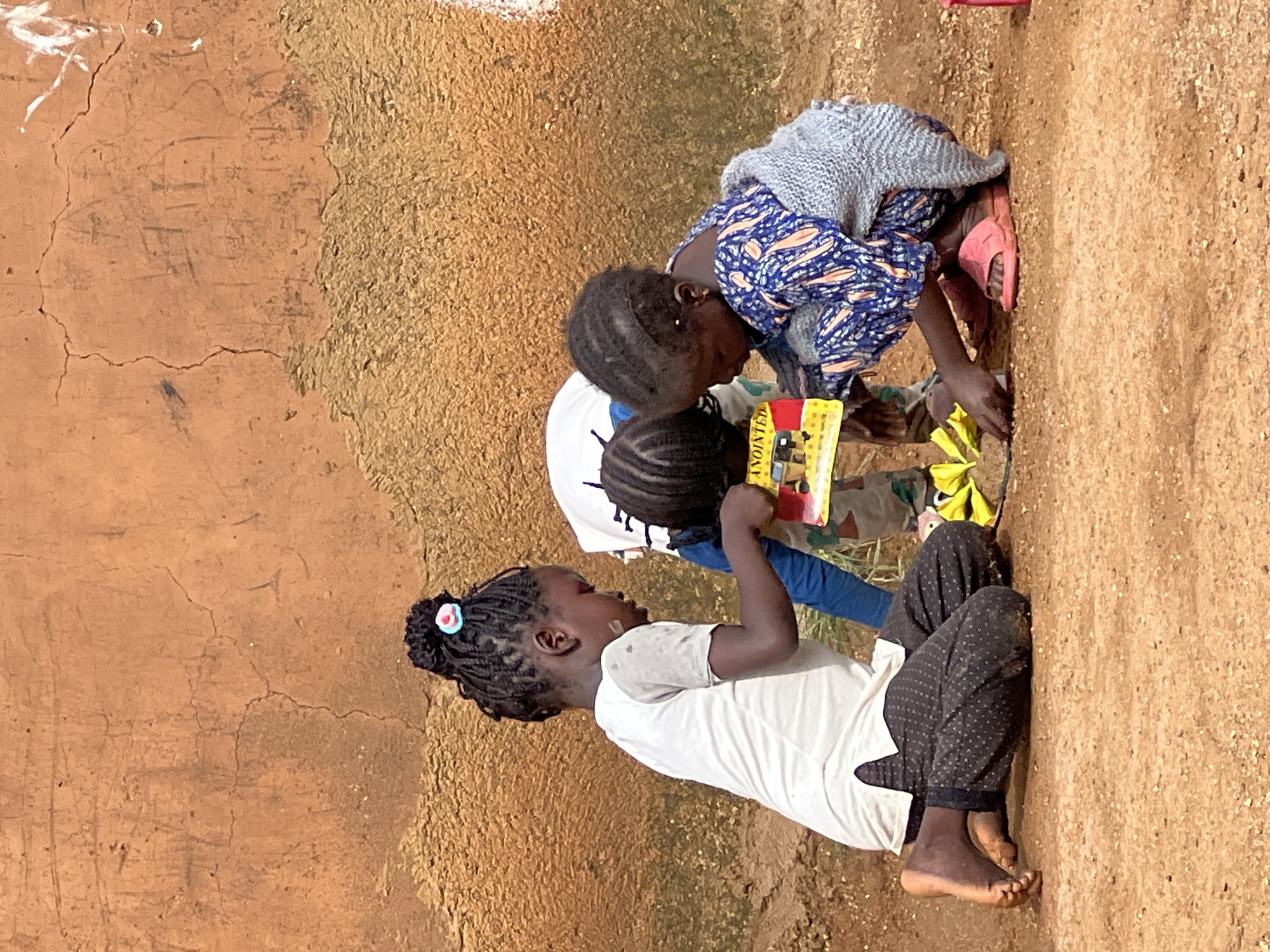 Three young African children are engaged in an activity on the ground in front of a textured, ochre-colored wall. The child on the left is kneeling on her right knee, wearing a white t-shirt and dark patterned pants. She is looking down at a yellow object. The middle child is also kneeling and is mostly obscured by the other two, with only their head and braids visible. The child on the right is crouching, wearing a blue and orange patterned dress and a grey knitted shawl draped over their shoulders. They are holding a small, rectangular object with yellow and red elements, which appears to be a book or a toy. A few yellow, leafy objects are scattered on the ground between the children. The ground is composed of reddish-brown earth with small pebbles. The wall behind them is rough and shows signs of age and wear. This scene is located in Jos, Nigeria.