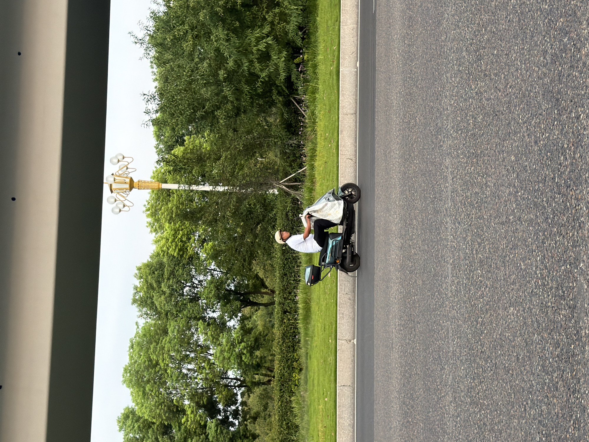 A person wearing a white t-shirt and black pants, and a light-colored helmet, rides a teal-colored electric scooter along a paved road. The scooter has a black storage box mounted on the back. The rider is carrying a white plastic bag, partially covering the scooter's front. The road is bordered by a concrete curb and a strip of grass, beyond which are trees and a decorative street lamp with multiple globes. The scene appears to be in the city of Jiangjin, China.