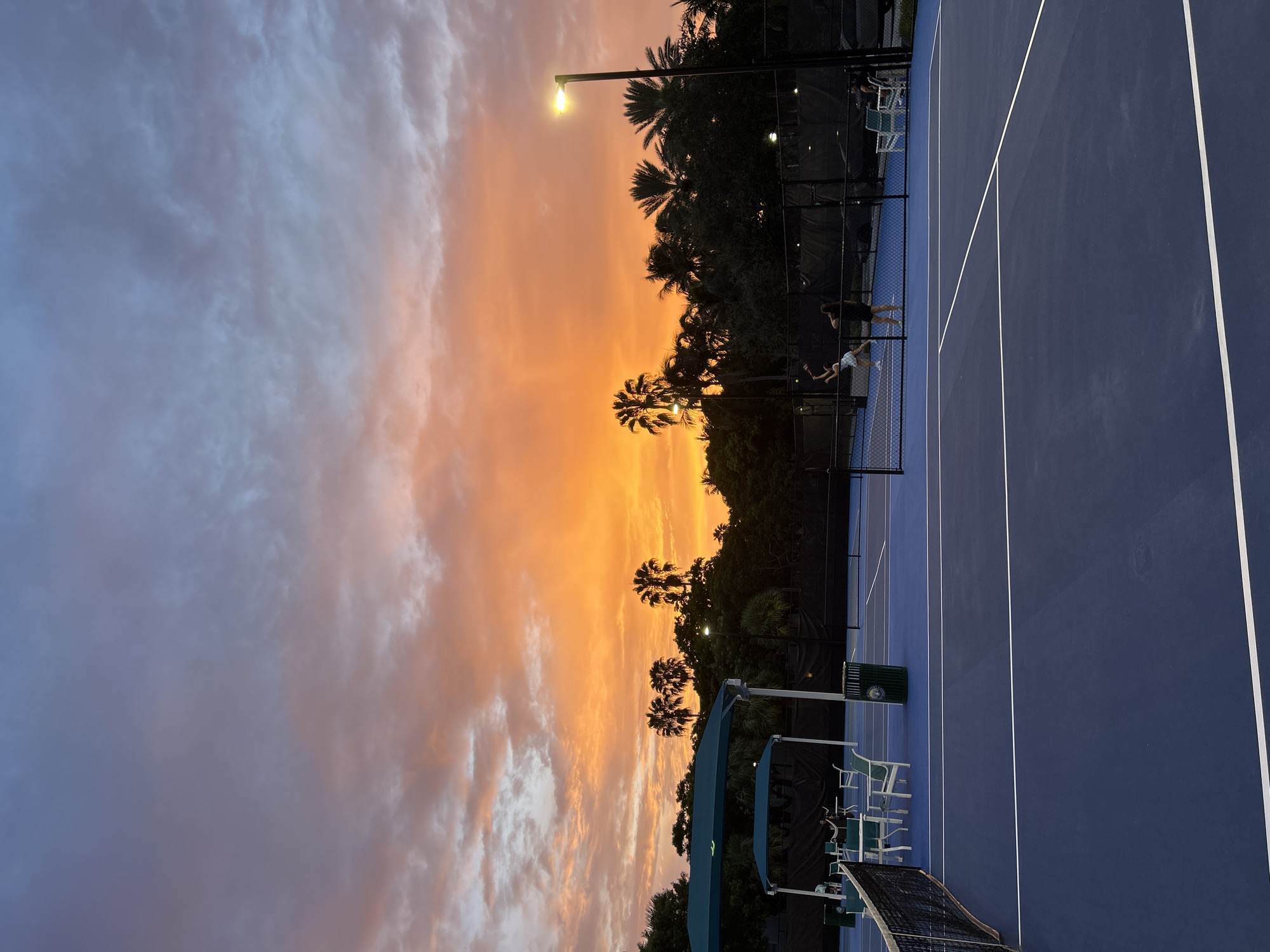 The image depicts a tennis court at dusk, with a vibrant orange and pink sunset painting the sky. Two individuals are engaged in a tennis match on the court, with one in mid-air seemingly hitting a ball. Palm trees and lush green foliage form a backdrop against the colorful sky. A tennis net divides the court, and to the left, there are shaded seating areas with tables and chairs. Streetlights are visible, illuminating parts of the court. The scene is set in Aventura, Florida, United States of America.