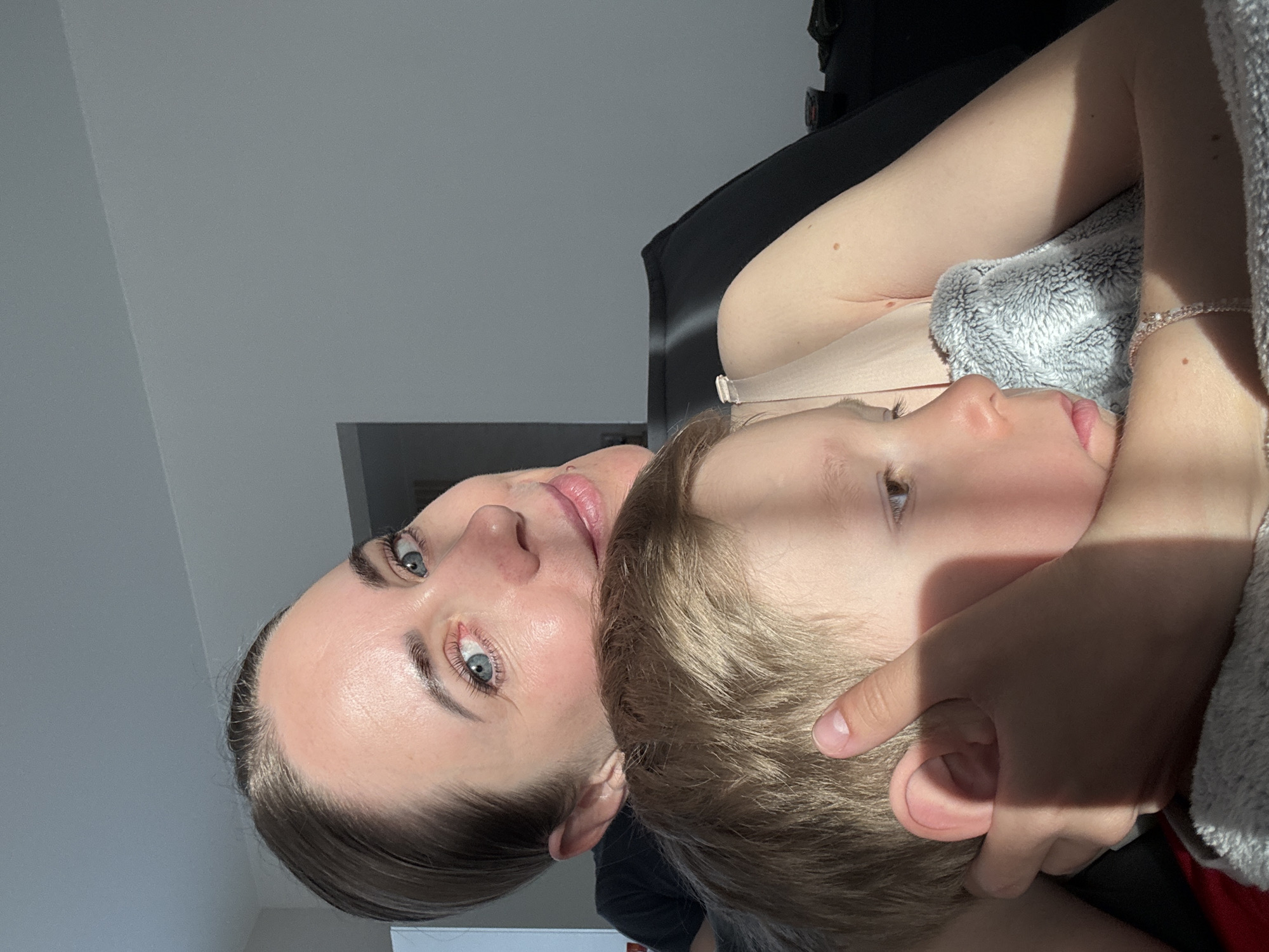 A woman and a child are in an embrace, with the woman's face visible and the child's head resting on her. The woman has fair skin, blue eyes, and her hair is pulled back. The child has blonde hair and fair skin. The background consists of a white wall and some dark objects. The lighting suggests it is daytime indoors. The location cannot be determined as Chaikovsky, Russia from the visual data available.