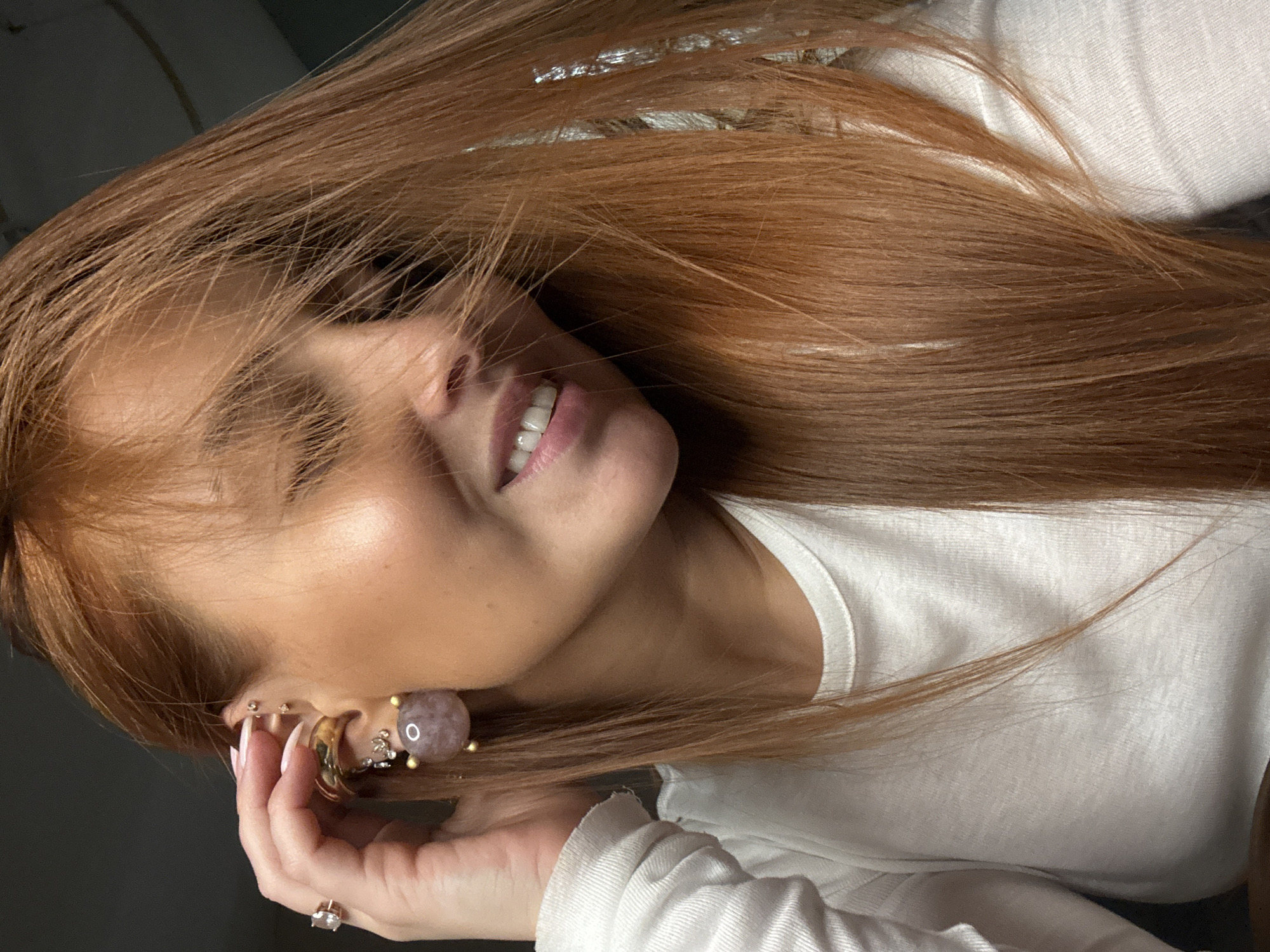 The image presents a close-up portrait of a person, likely female, with long, flowing reddish-brown hair cascading around her face and across a white long-sleeved top. Her head is tilted to the side, and she is smiling, revealing white teeth, which conveys a sense of happiness or contentment.
The subject's left ear (viewer's right) is prominent, adorned with several earrings, including a notable large, light purplish-pink spherical earring with gold-colored accents. Smaller gold-toned hoops and studs are also visible on both ears. One hand, featuring manicured nails painted in a light, natural shade and a ring with a light-colored stone, is gently positioned near her ear, possibly resting or adjusting her hair or jewelry.
The background is dark and out of focus, indicating an indoor setting. The lighting is soft, creating highlights on her hair and skin. The overall scene suggests a casual and relaxed moment. There is no visible text in the image. The context for the image is Chaikovsky, Russia.